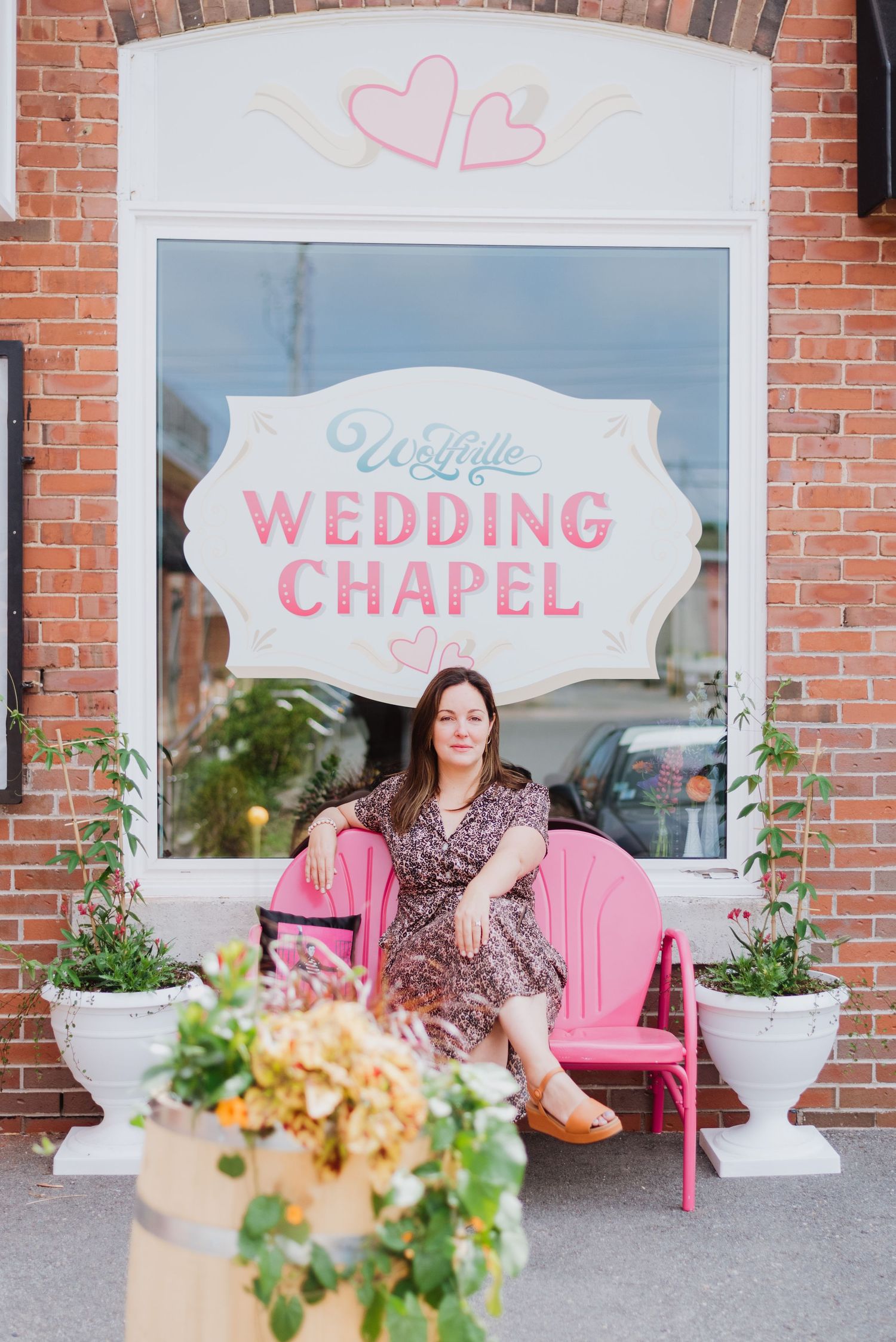 Woman in floral dress sitting on pink bench outside We Little Wedding Chapel with brick exterior and flower planters.