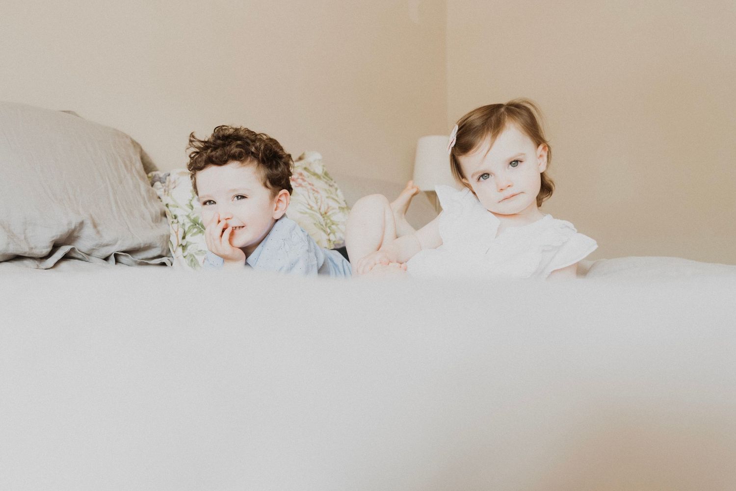 Two young toddlers peeking over a white bed, a boy and girl posing together in soft natural light.