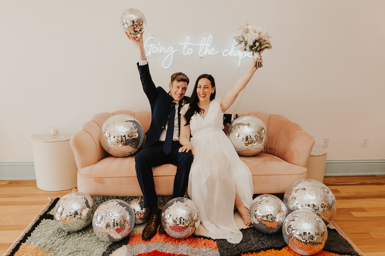 Newlyweds celebrate on a pink sofa surrounded by silver disco balls with a neon sign reading going to the chapel.