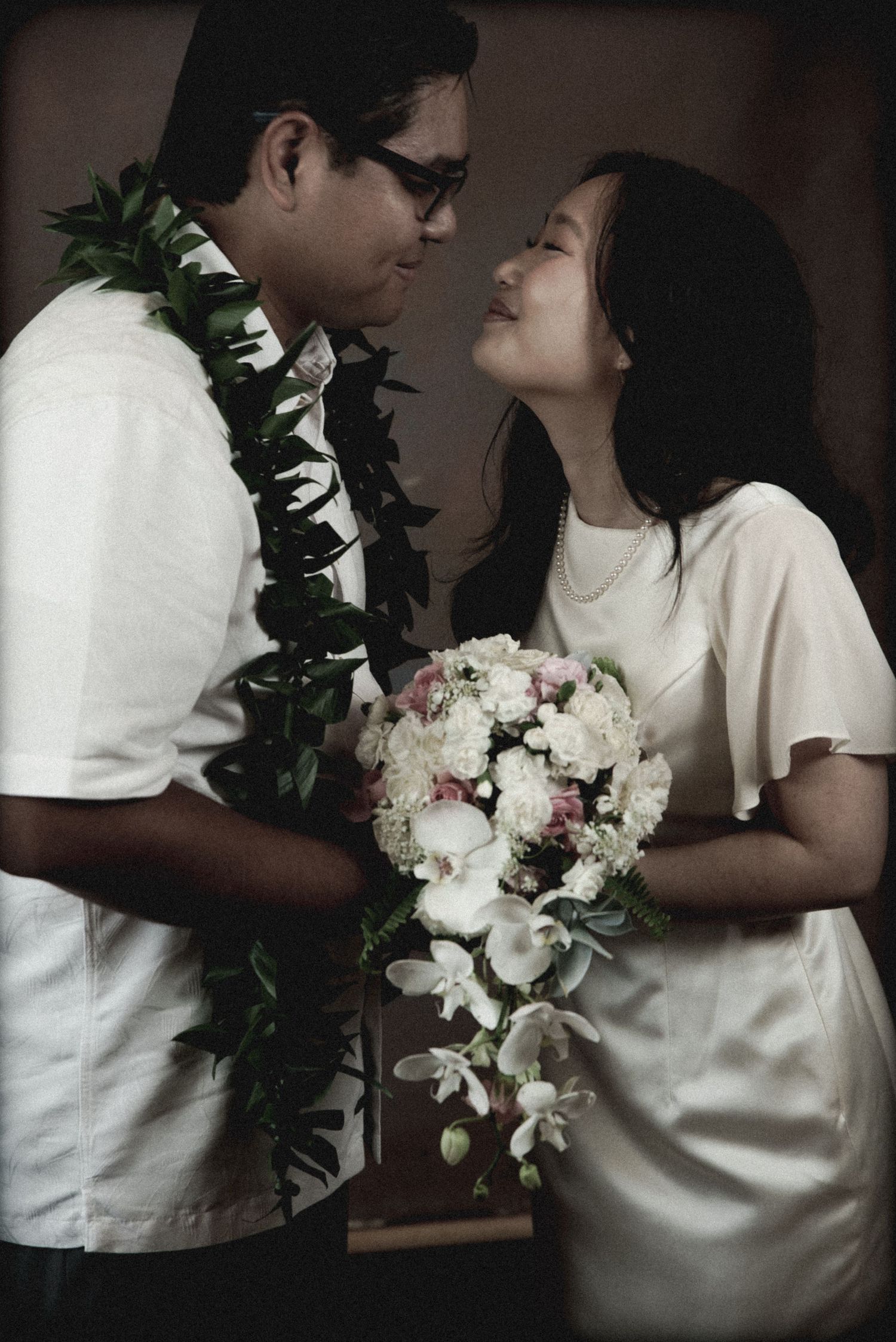 A romantic couple in white clothing shares an intimate moment with a floral lei and wedding bouquet.