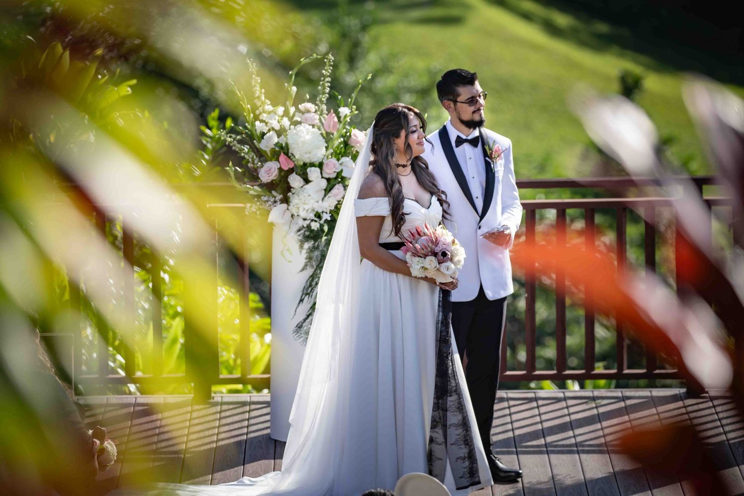 A couple in wedding attire shares a romantic moment on a bridge surrounded by soft green foliage.