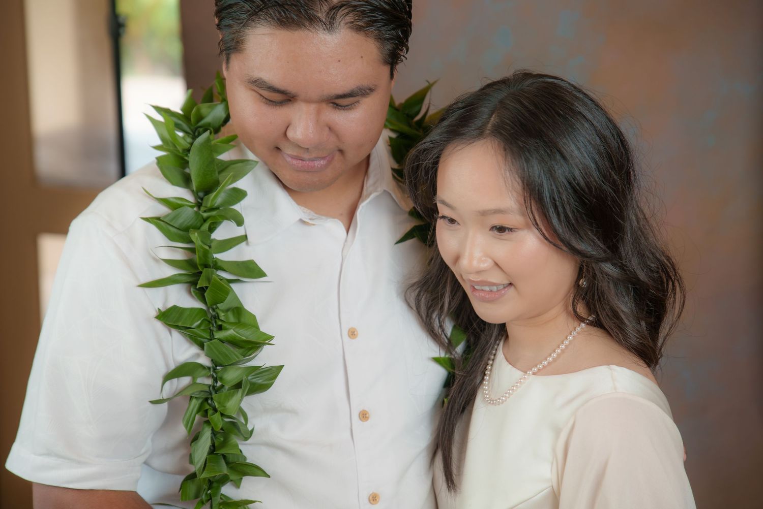 Couple in white attire smiling, man wearing green leaf lei, woman with pearl necklace, elegant outdoor setting.