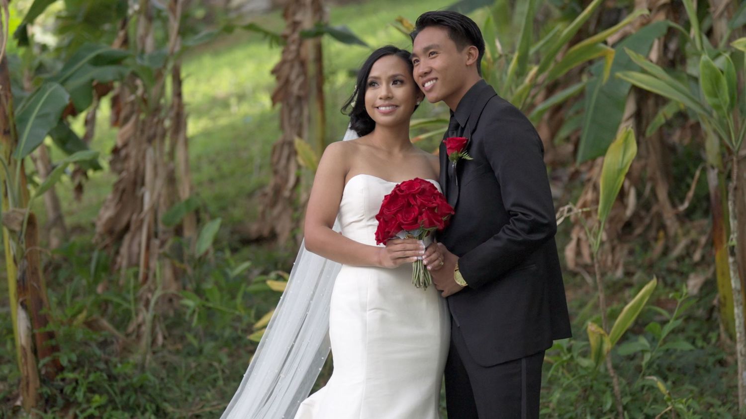 A bride in a white dress holds red roses while standing with her groom in a black suit near tropical foliage.