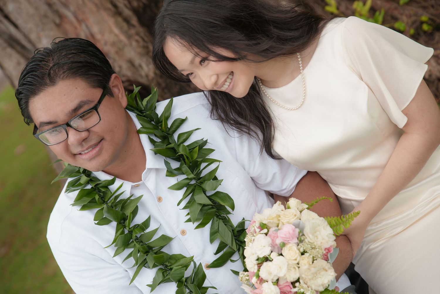 Couple in wedding attire with green lei and white bouquet smiling together outdoors in Hawaii.