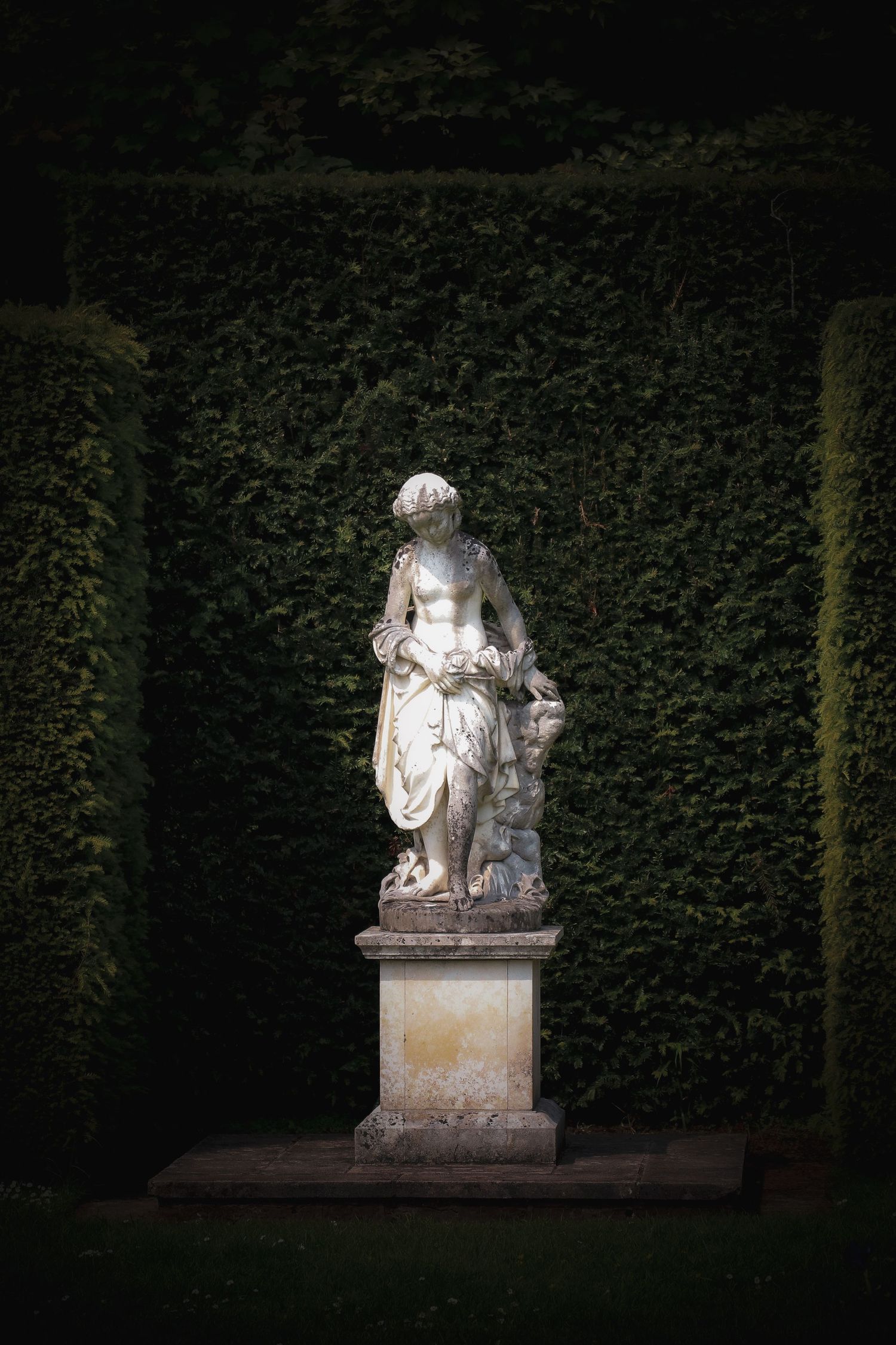 Classical white marble statue of a female figure on a pedestal surrounded by neatly trimmed dark hedges.