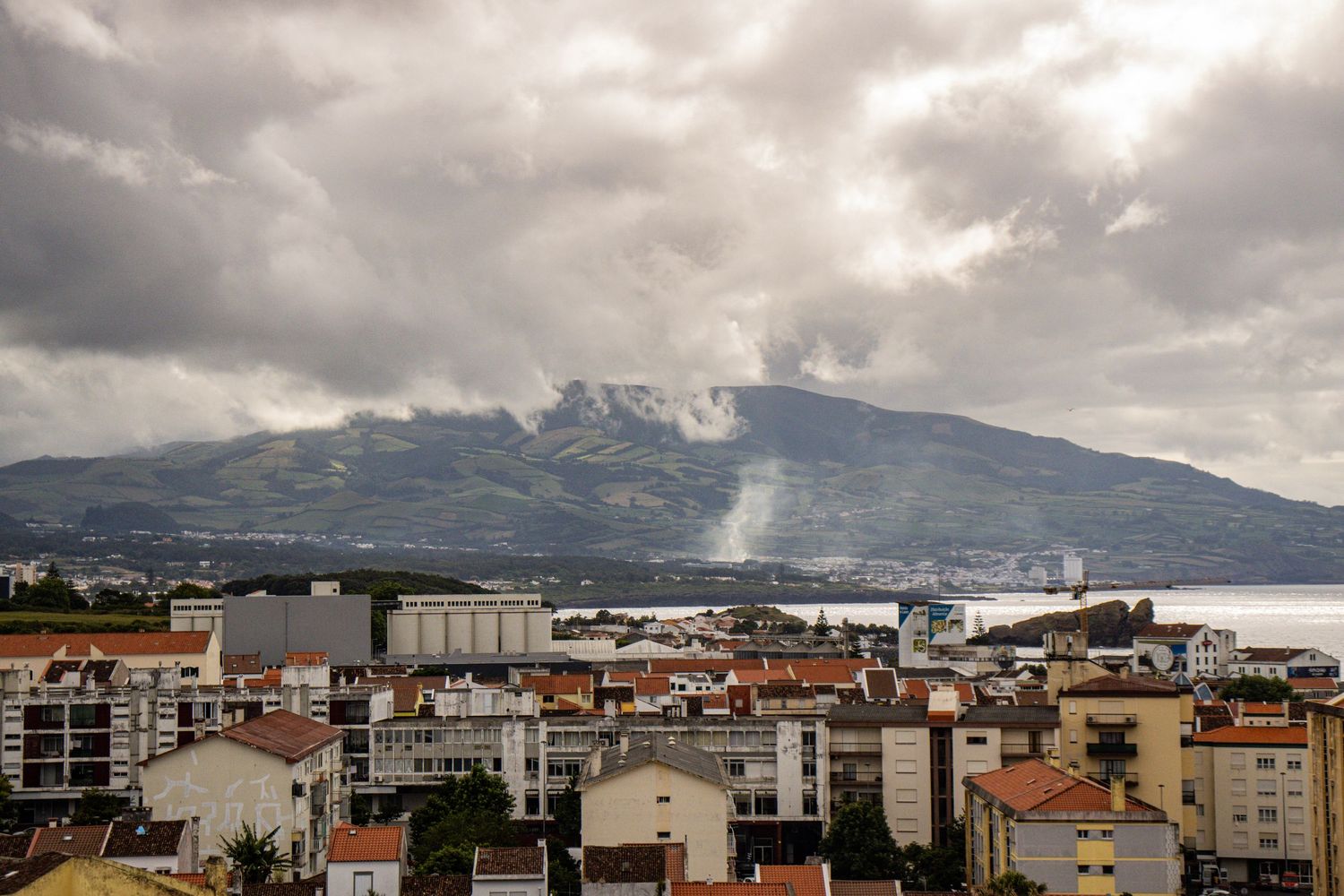 Panoramic view of a coastal city with terracotta rooftops beneath dramatic cloudy skies and a mountain backdrop.