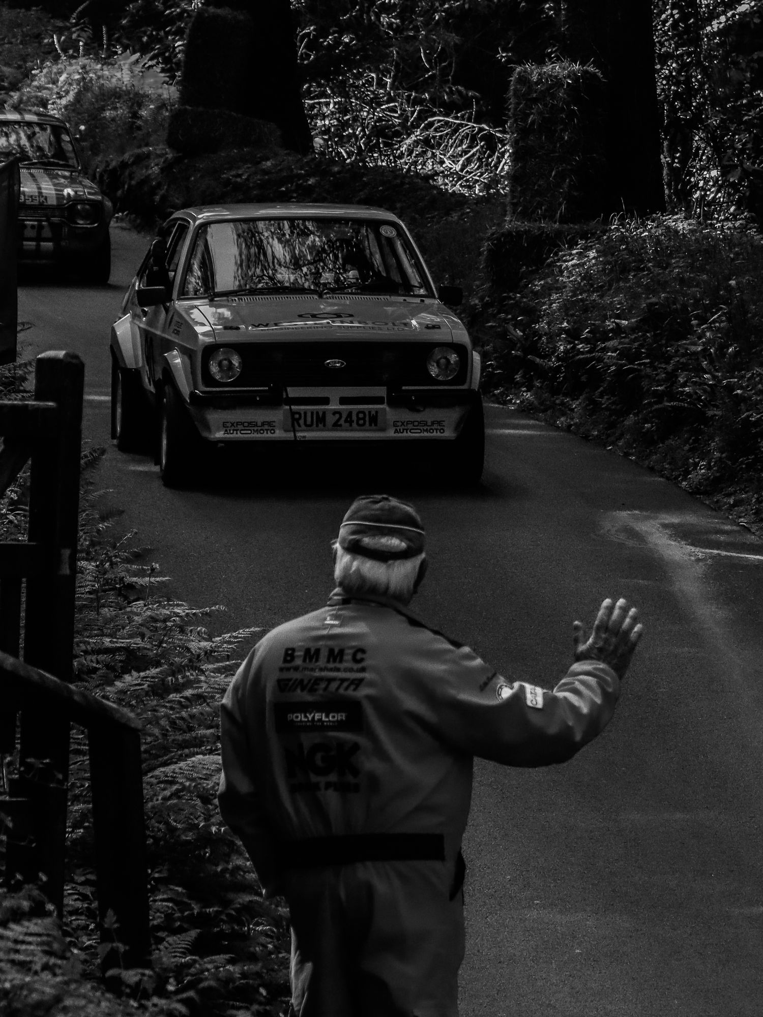 Black and white photo of a marshal waving a vintage Ford Escort rally car on a forest road.