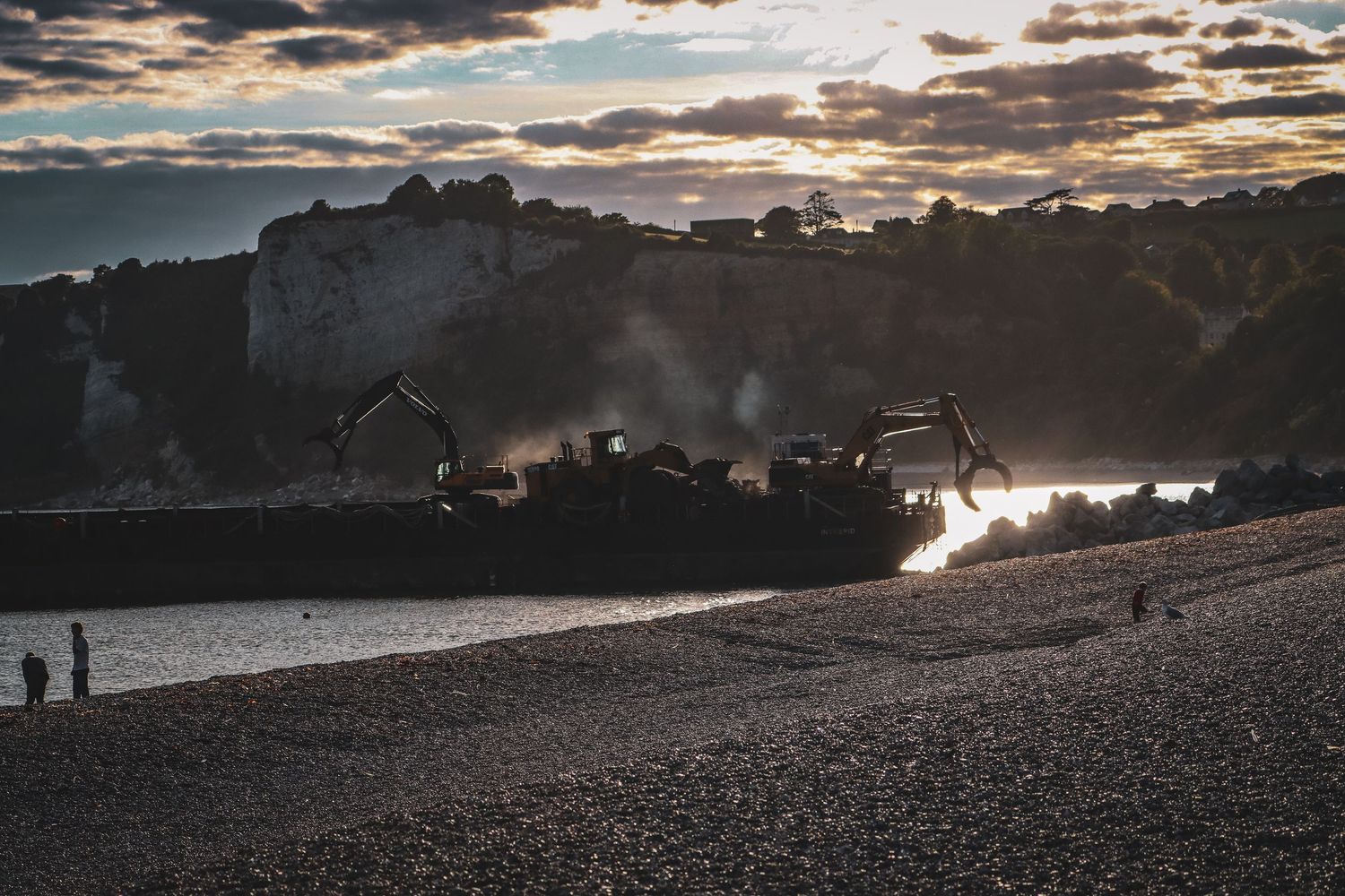 Silhouetted excavators working on a shingle beach at sunset with dramatic cloudy skies.