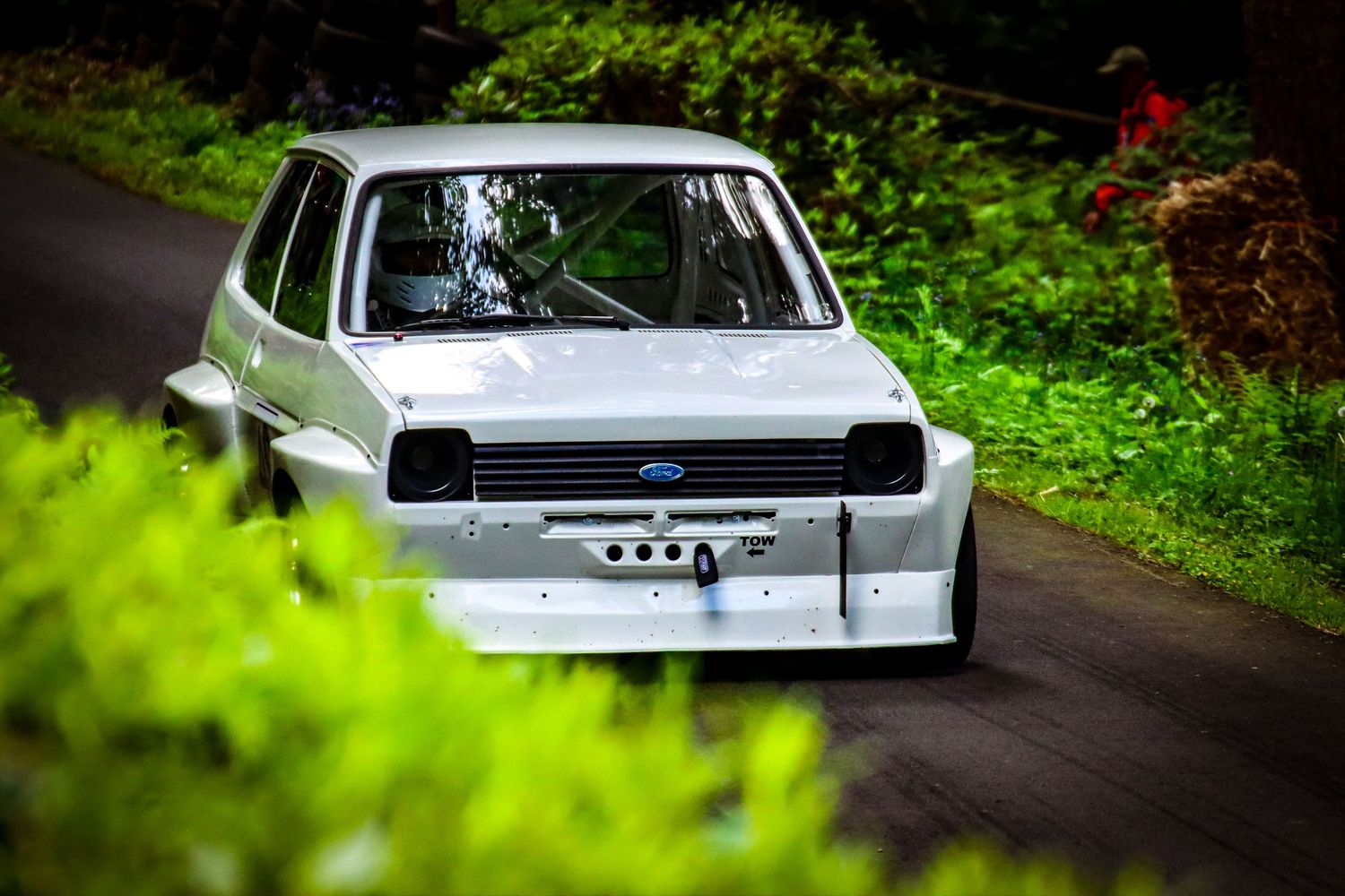 White vintage Ford Fiesta race car on lush green tree-lined track.