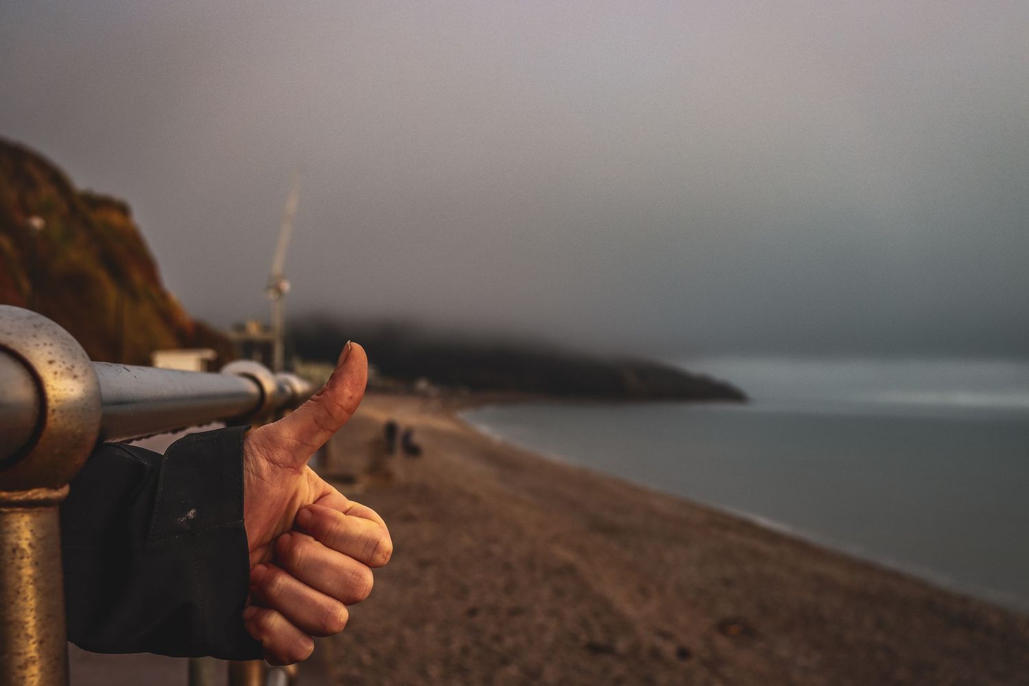 Person giving thumbs up near coin-operated telescope on foggy beach coastline.