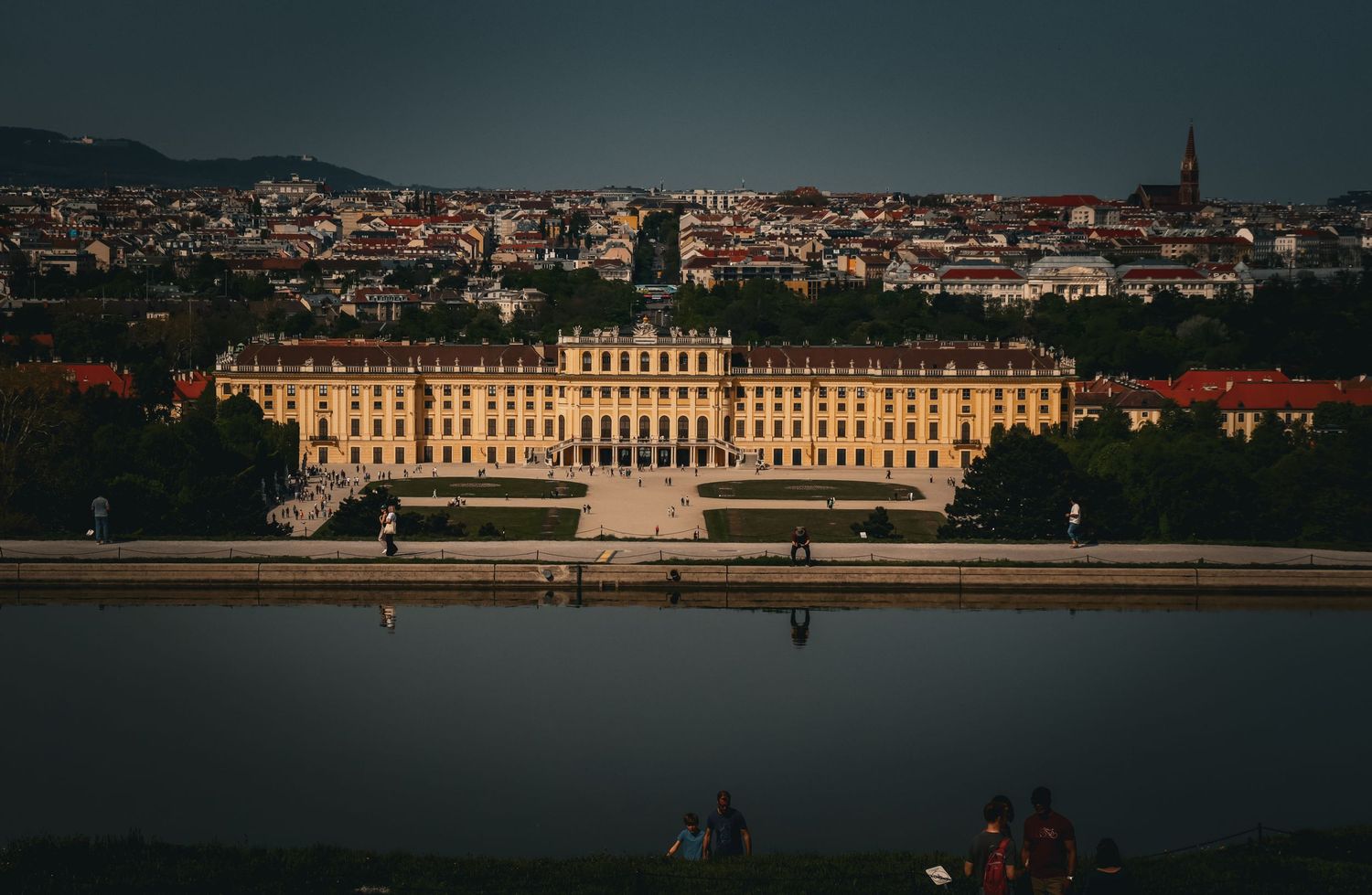 The grand Schonbrunn Palace in Vienna glows at dusk, reflected in a nearby pool with city views beyond.