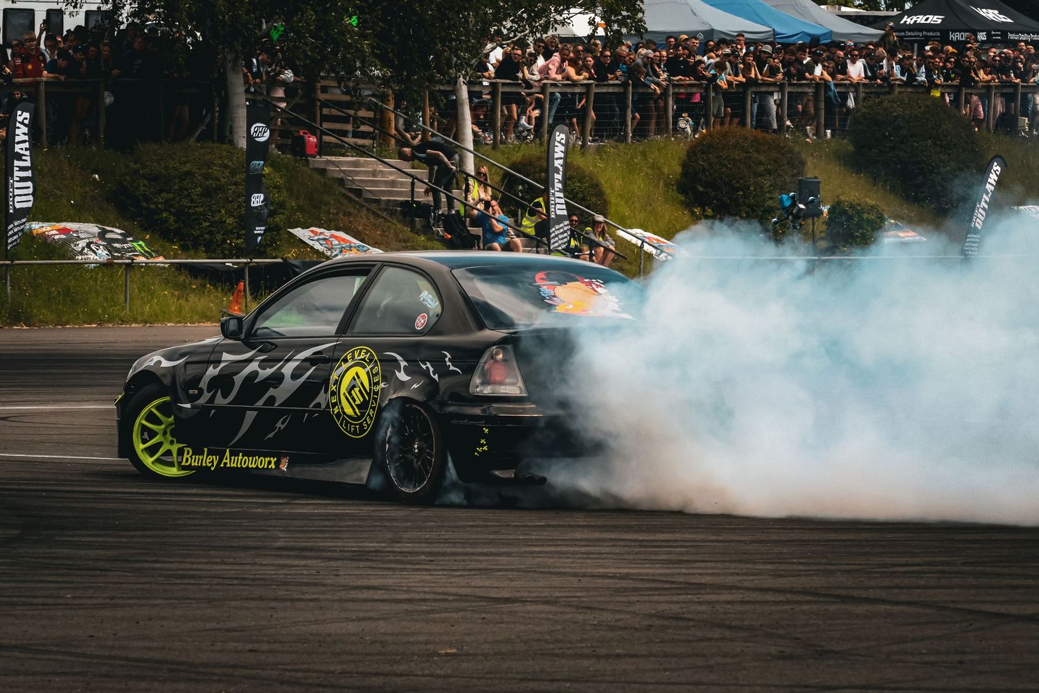 Black drift car performs smoky burnout on race track surrounded by spectators.