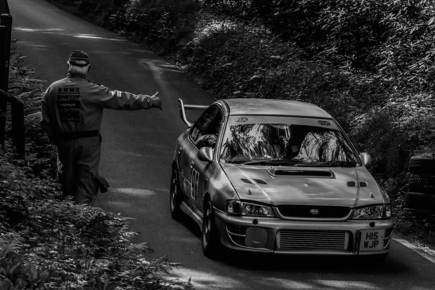 Black and white rally car racing on a narrow forest road with a marshal signaling nearby.