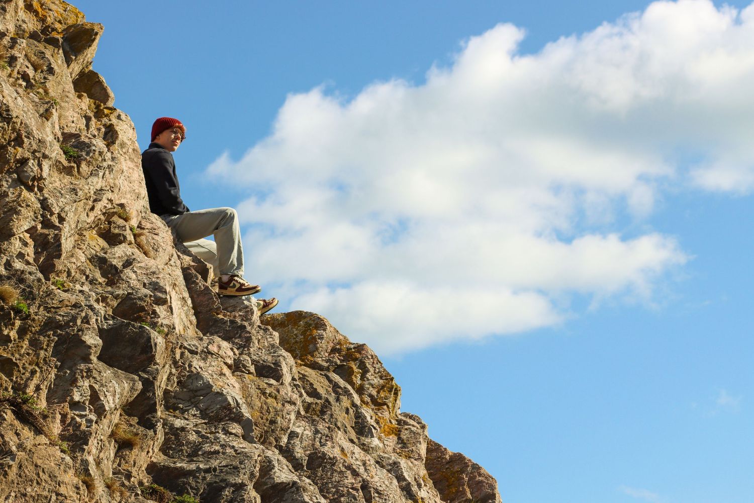 A person with red hair sitting on rugged rocky cliff face against a bright blue cloudy sky.