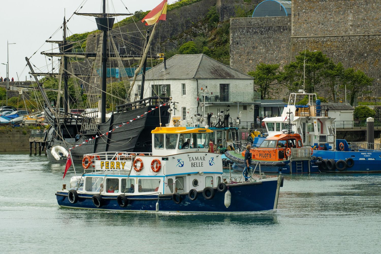 A blue and white ferry boat navigating a harbor with tall ships and buildings in the background.