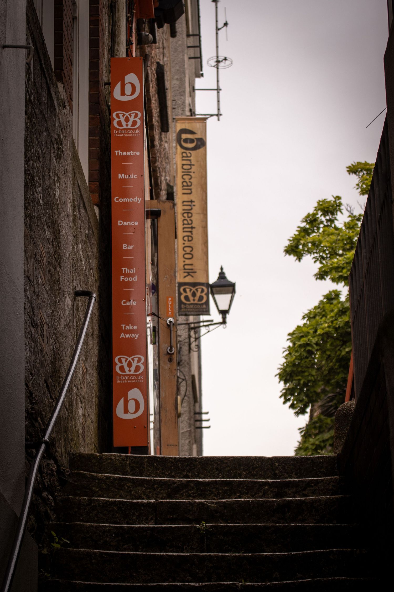 Narrow alley steps leading to Barbican Theatre with red banners listing theatre, music, comedy, dance, bar, and cafe.