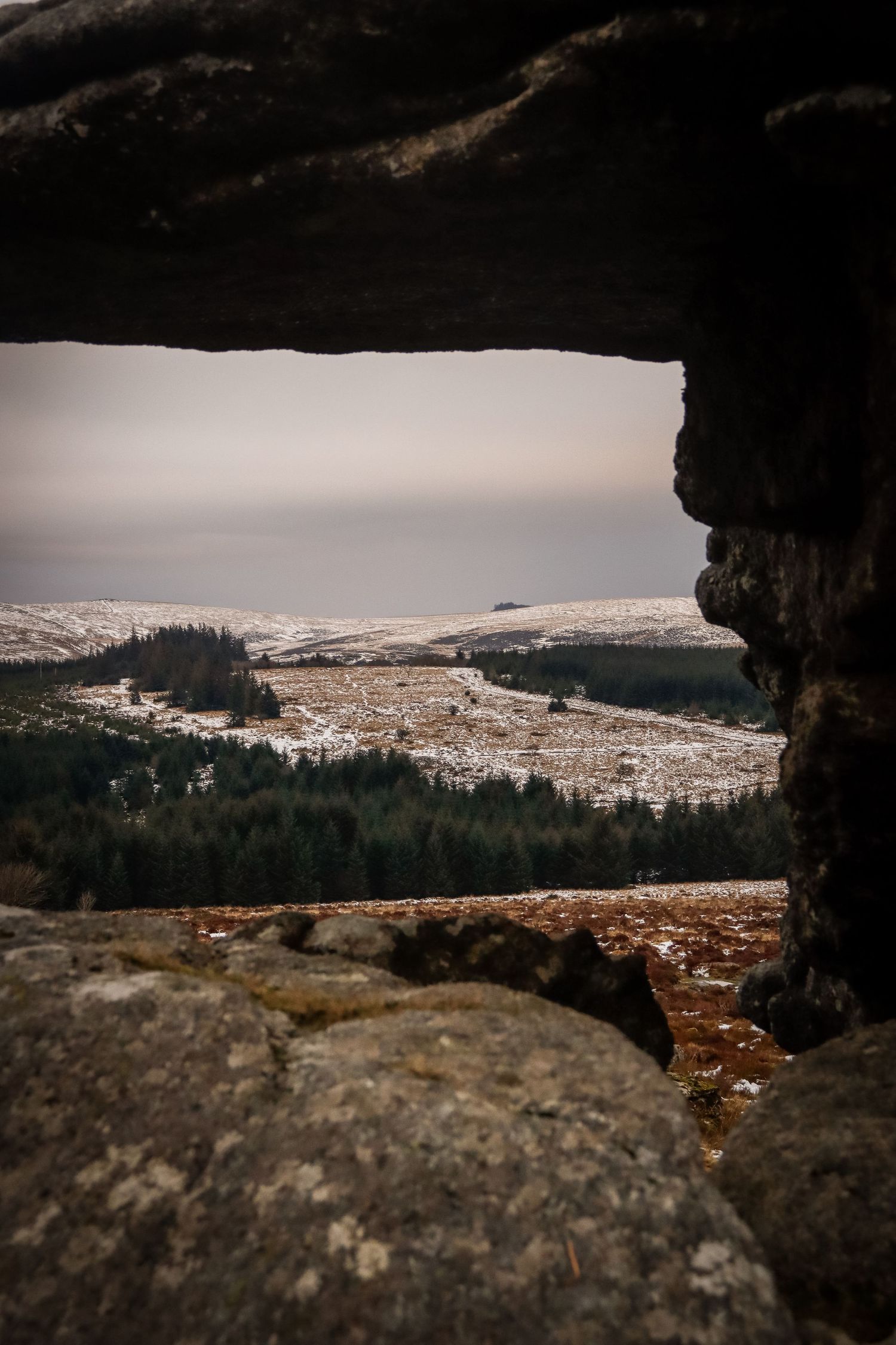 Snow-dusted moorland landscape viewed through a natural rock frame opening on a cloudy day.
