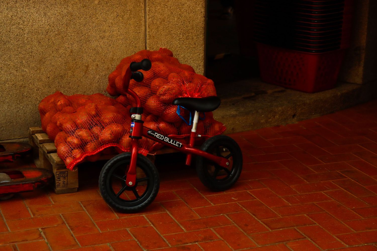 Red mesh bags of oranges stacked on a small red balance bike outside a building on brick pavement.
