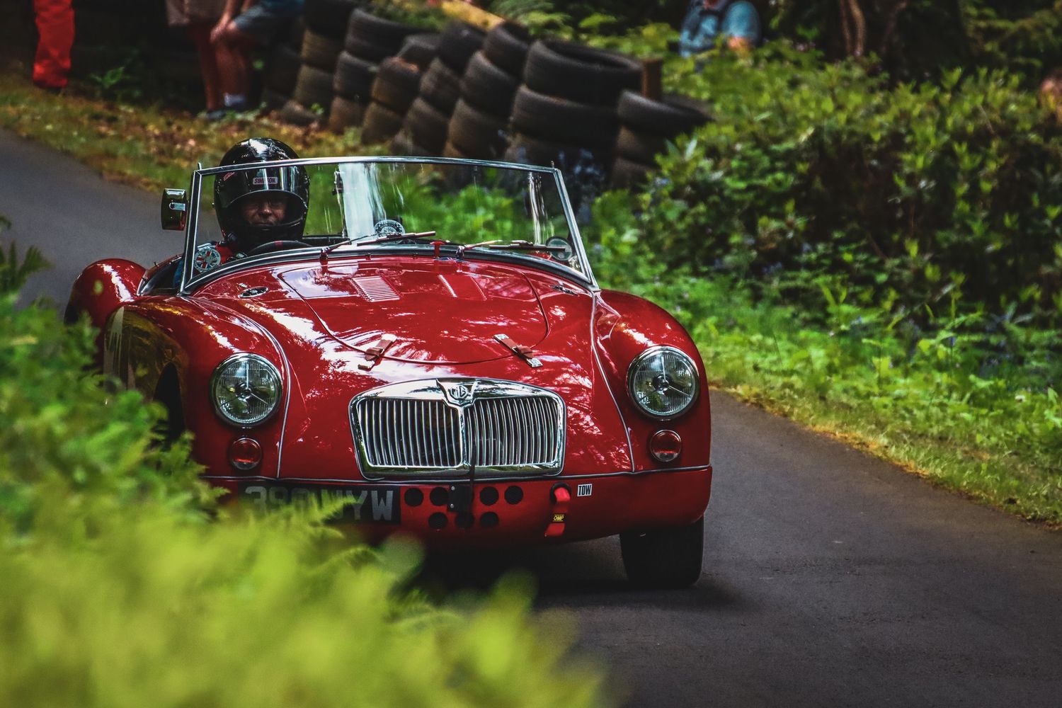 Red classic MGA convertible sports car racing on green tree-lined circuit.