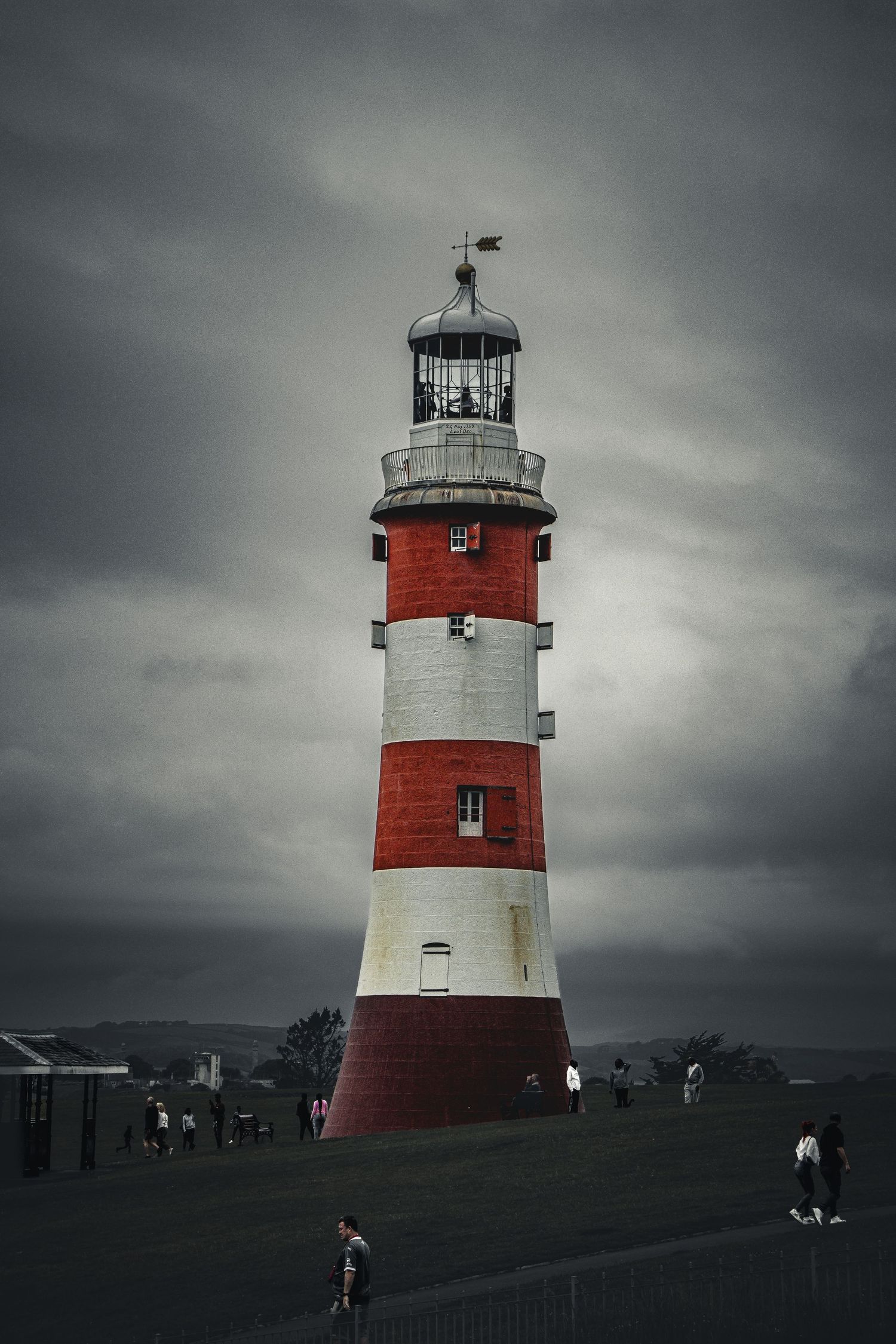 Red and white striped lighthouse under dramatic stormy grey skies with visitors nearby.