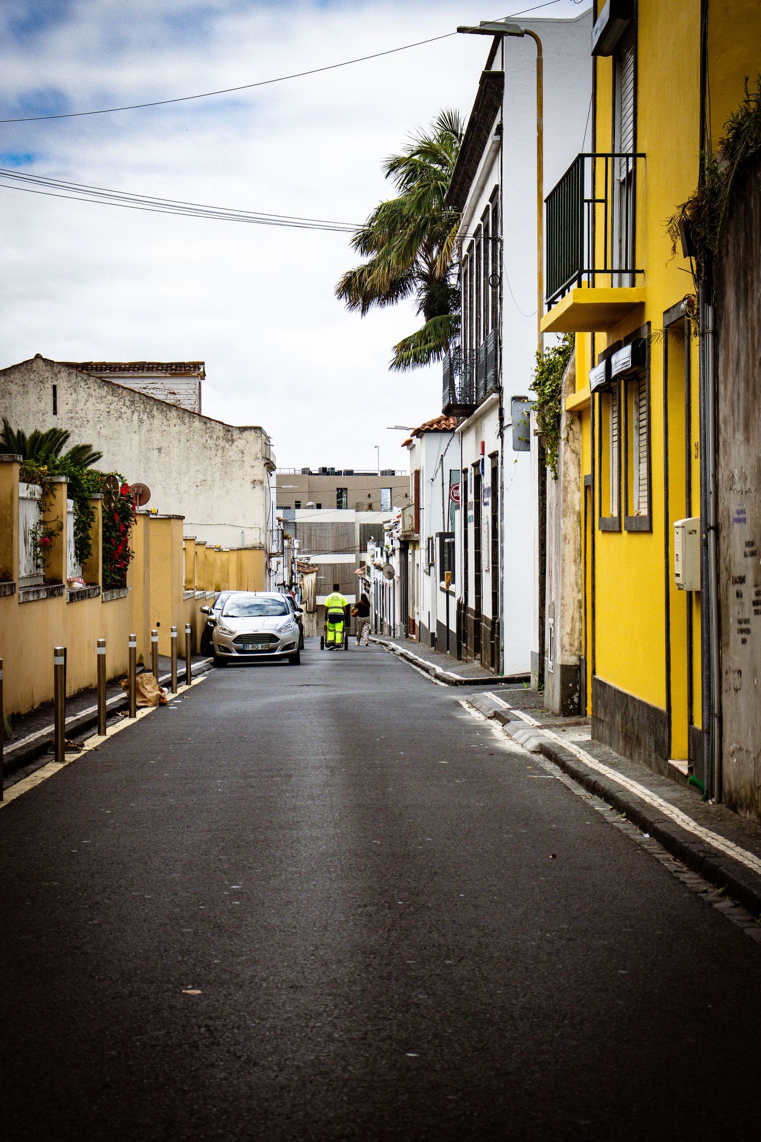 Narrow street in a Spanish town lined with yellow and white buildings, a parked car, and a worker in hi-vis.