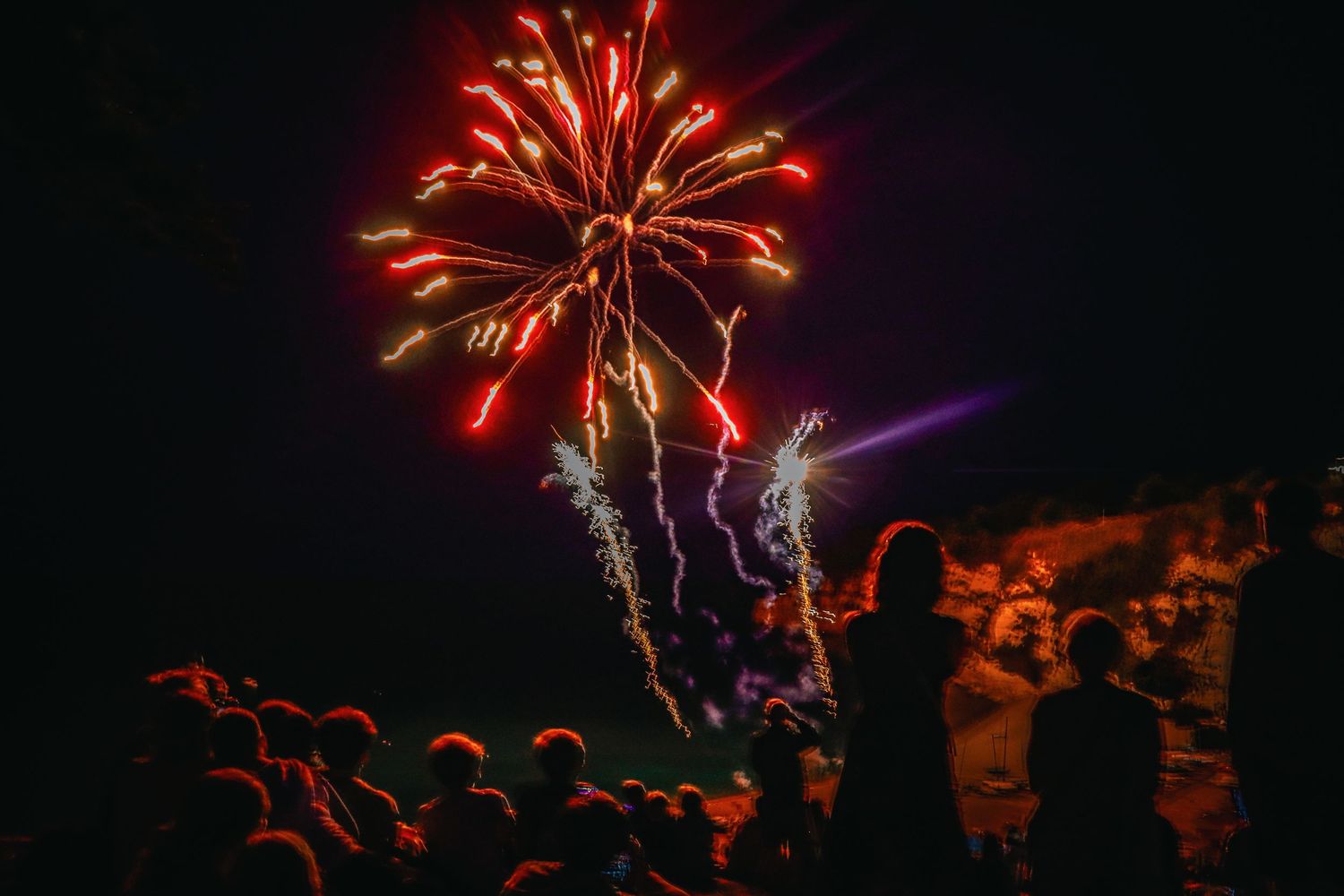Crowd of silhouetted spectators watching vibrant red and gold fireworks exploding in a dark night sky.