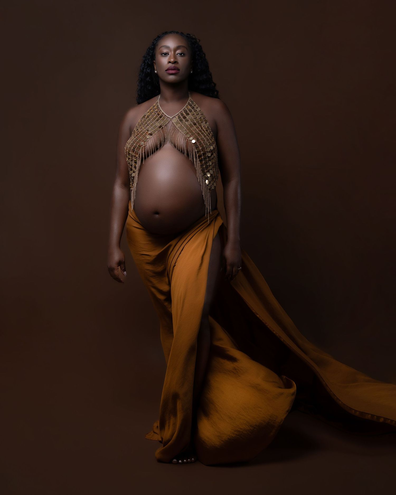 Pregnant woman in golden drape and beaded top poses elegantly against a dark brown studio background.