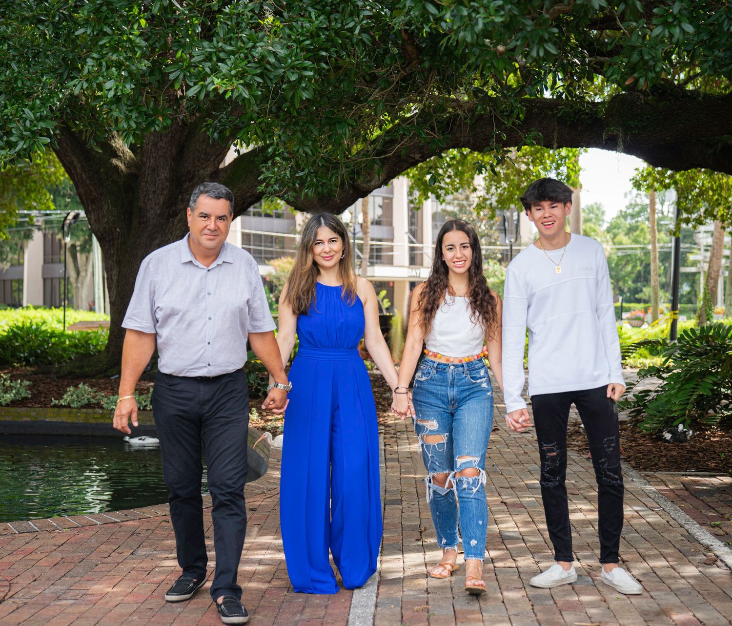 Group of people standing together on a brick path near a pond with trees and greenery in the background for a family photograph.