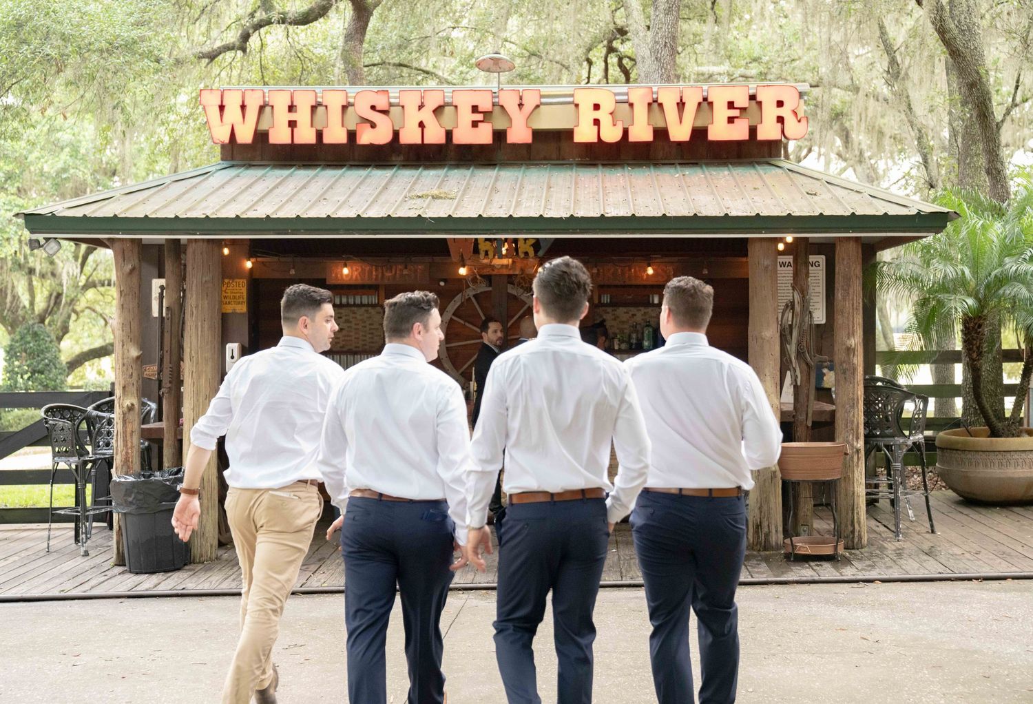 Four people wearing formal attire stand arm in arm under a rustic wooden sign that reads 'Whiskey River'.
