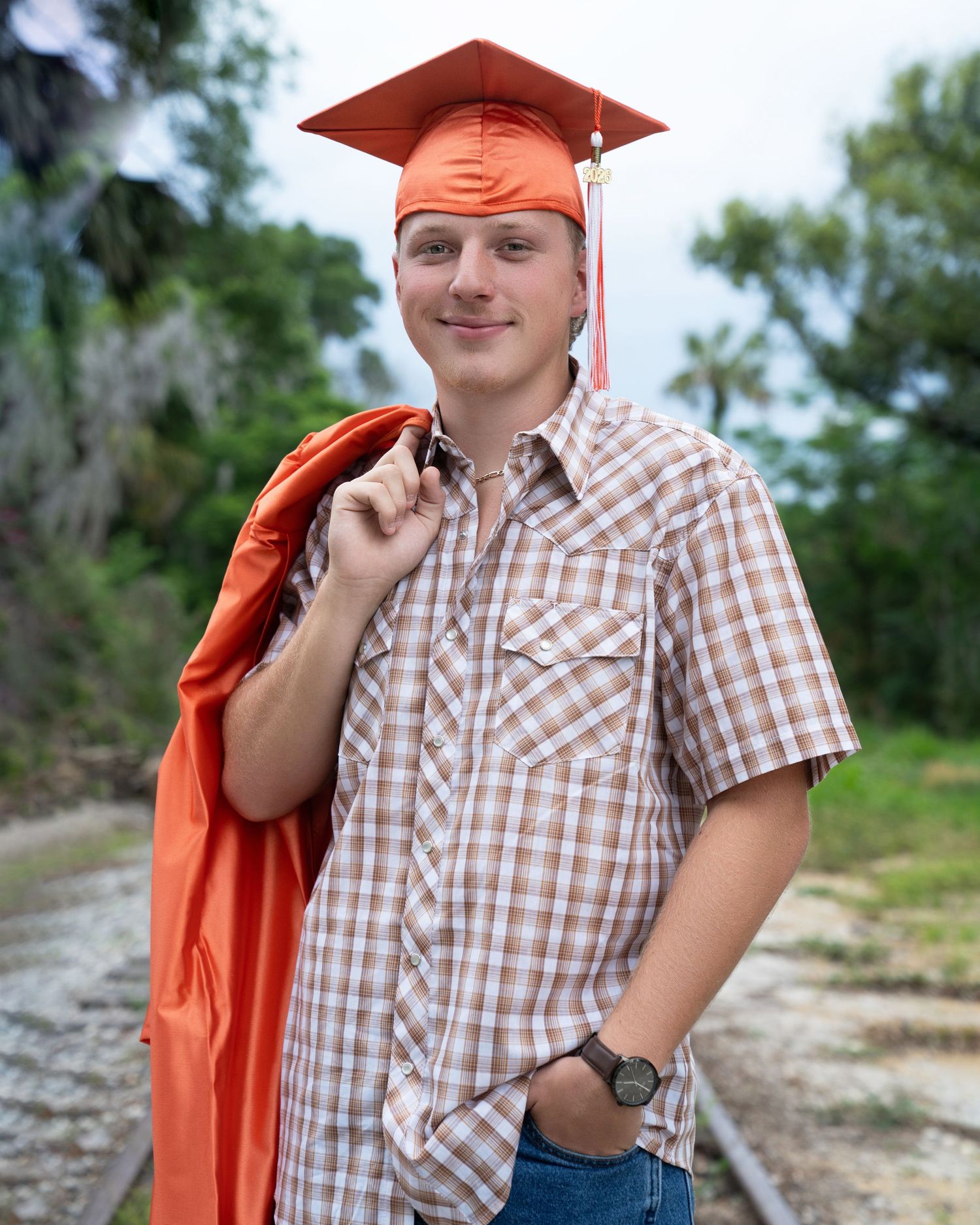 Young man in orange graduation cap and gown poses on railroad tracks, smiling in plaid shirt and jeans.