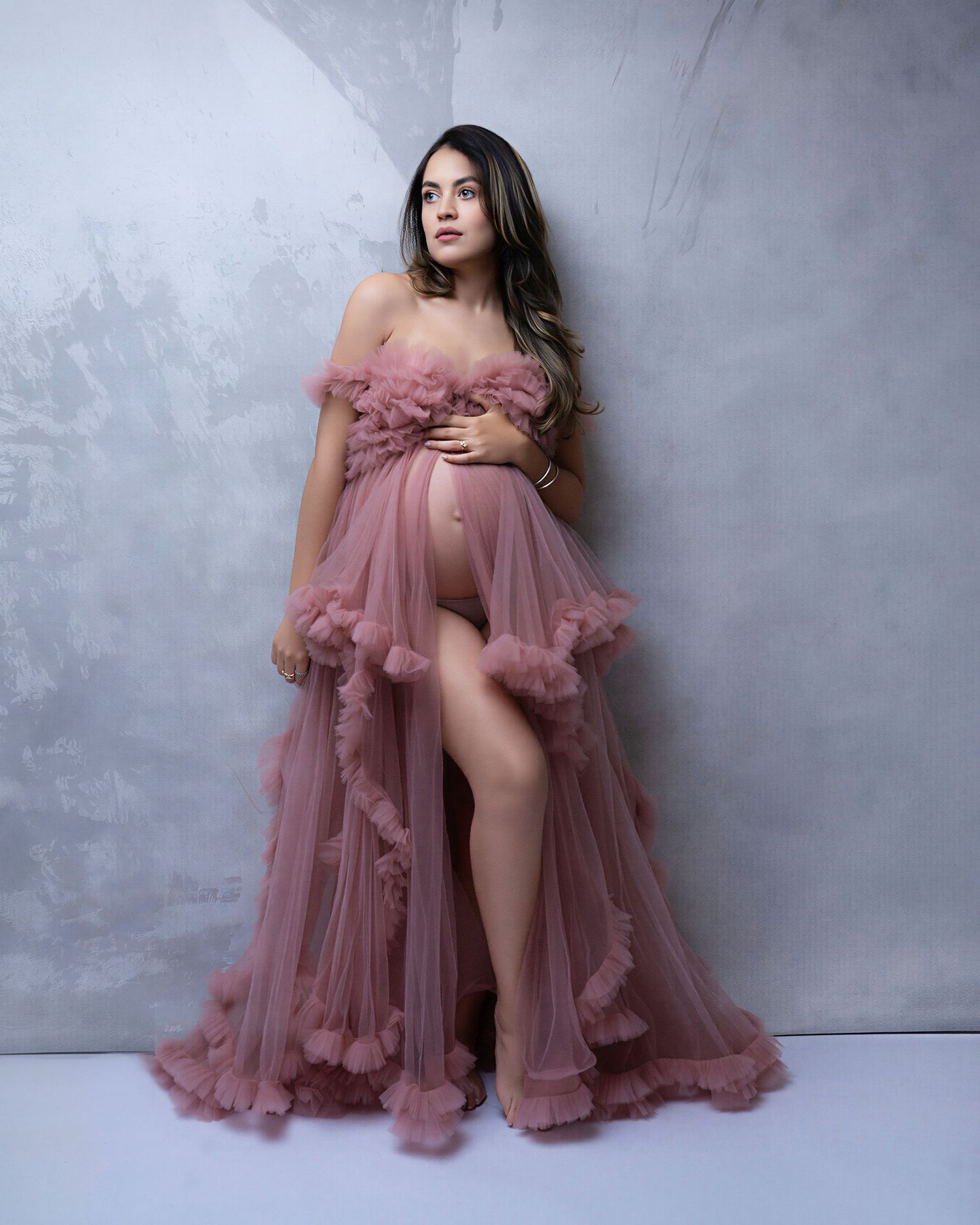 Pregnant woman in elegant dusty rose tulle maternity gown with ruffles, posing gracefully against gray backdrop.