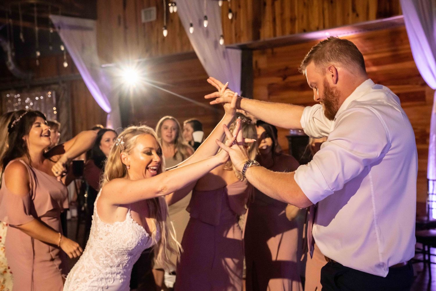 Wedding reception guests dance and celebrate on a dimly lit dance floor with purple lighting effects.