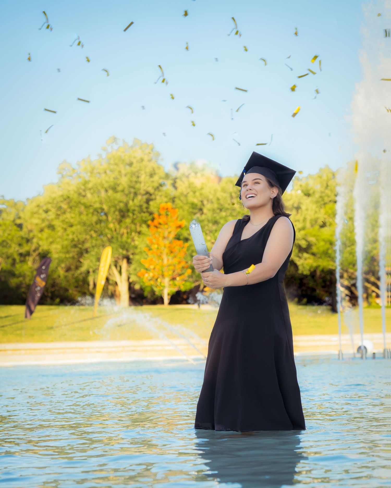 Graduate in black cap and gown celebrates in water with confetti falling.
