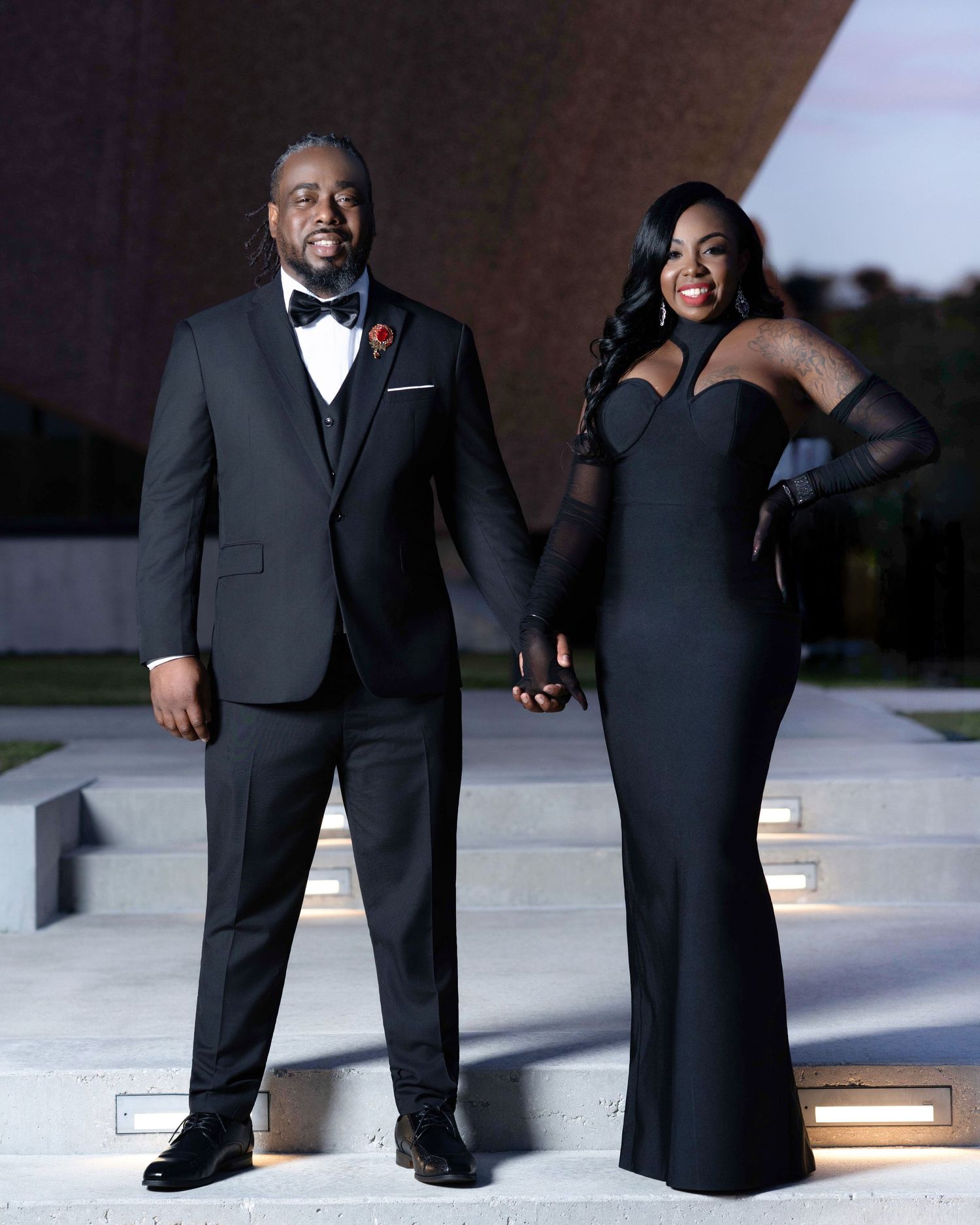 A couple in formal black attire shares a joyful moment while posing outdoors for wedding photos.