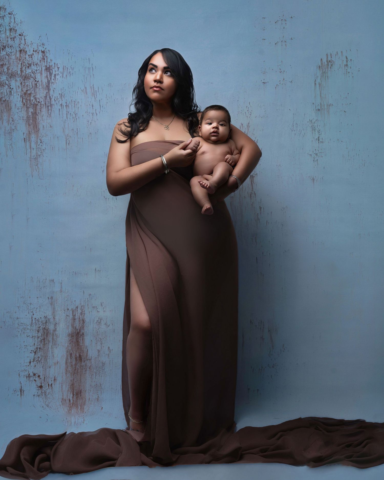 Mother and baby pose in brown flowing dress against textured blue wall.