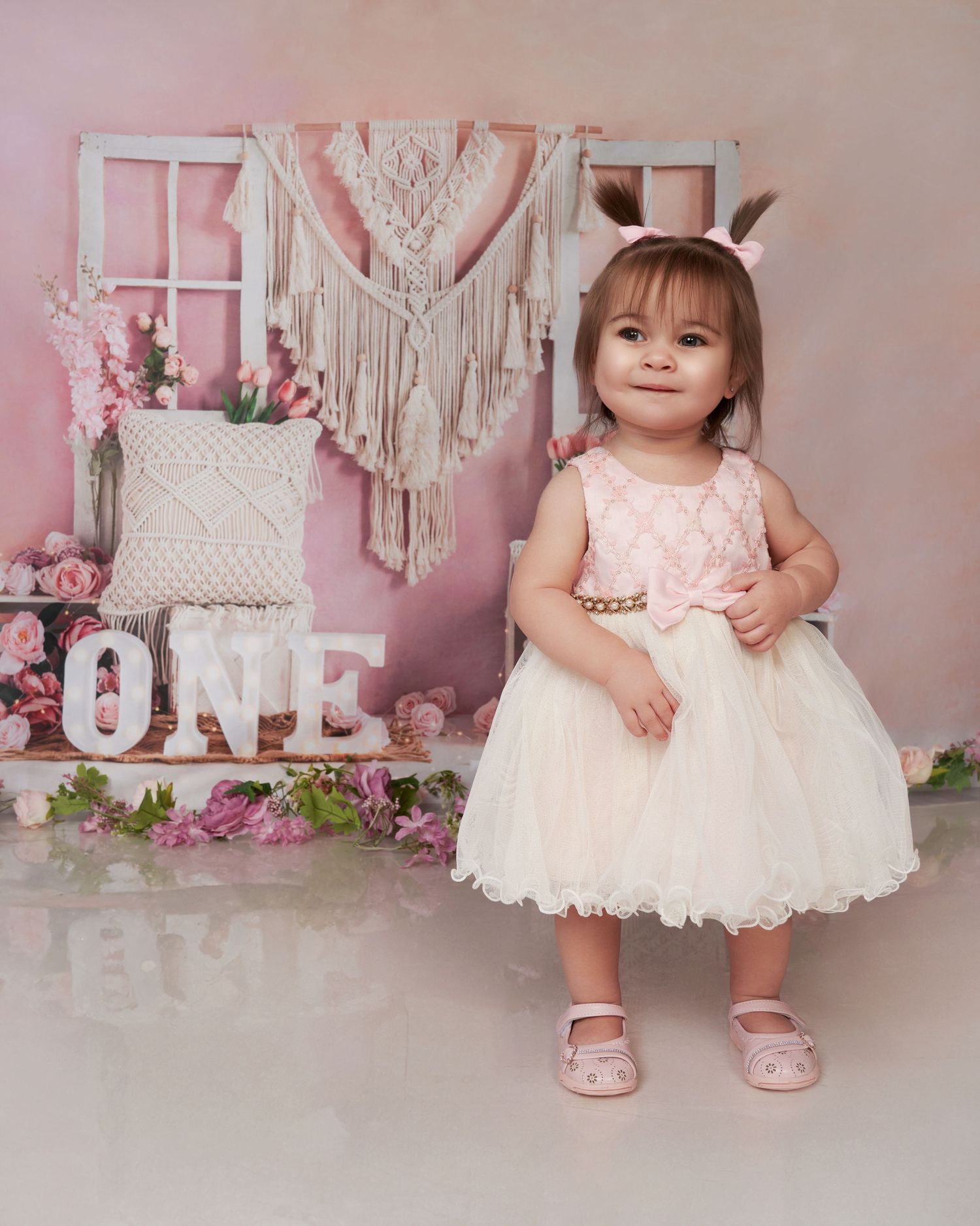 Baby in white lace dress poses for first birthday photos with pink decorative backdrop.