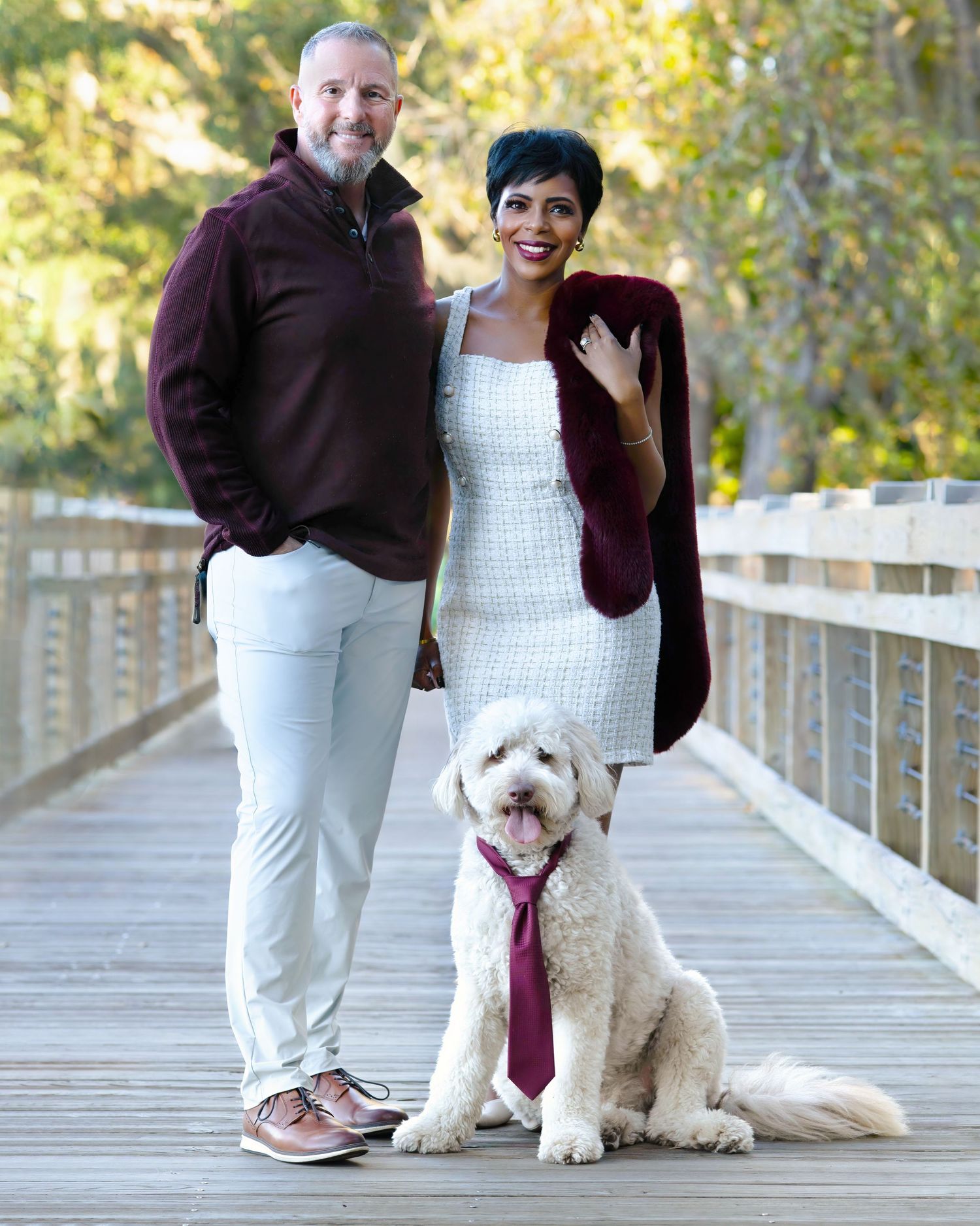 A couple poses with their fluffy white doodle dog wearing a matching burgundy tie on a wooden bridge.