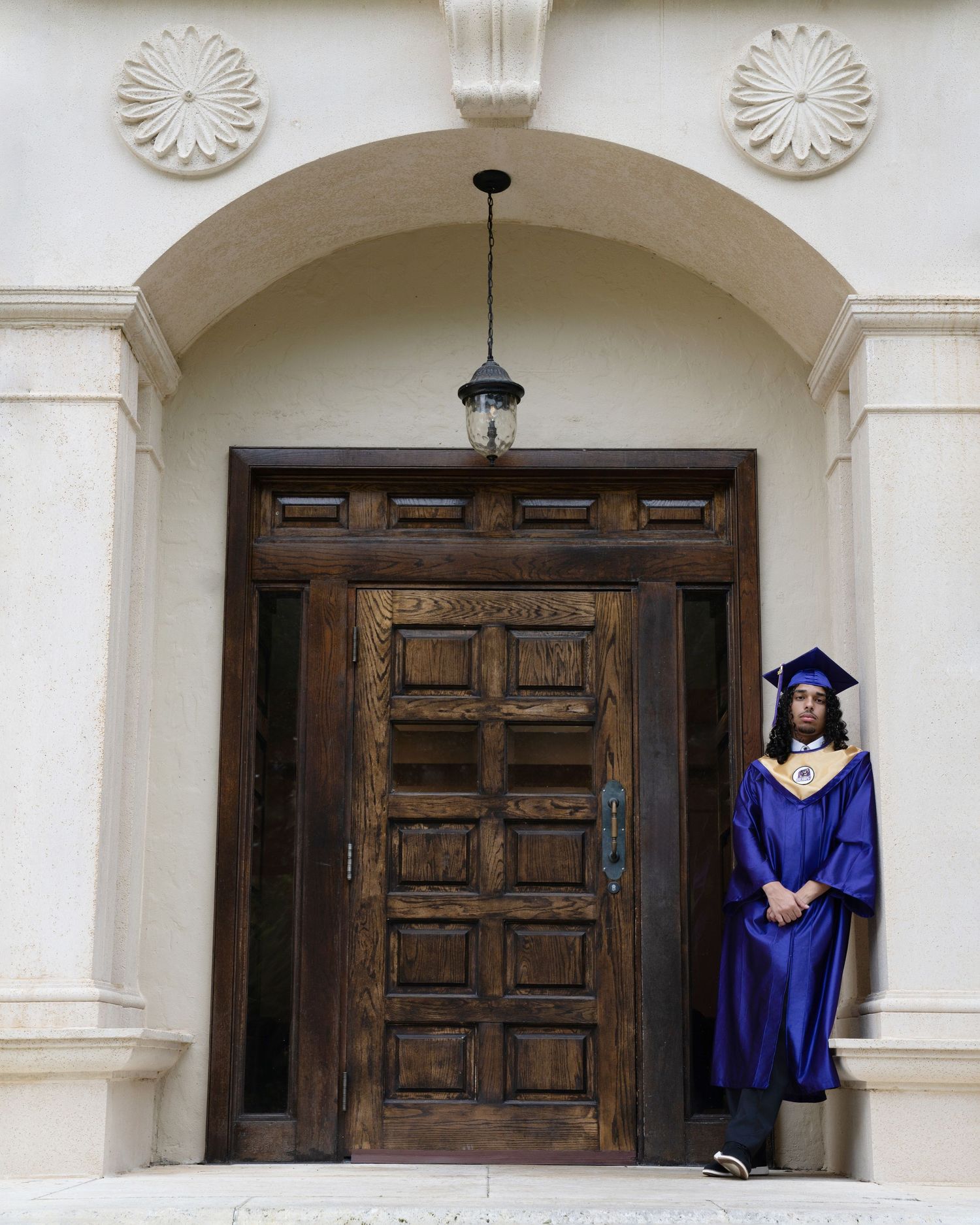 Graduate in blue cap and gown stands proudly before ornate wooden doors of a classical building.