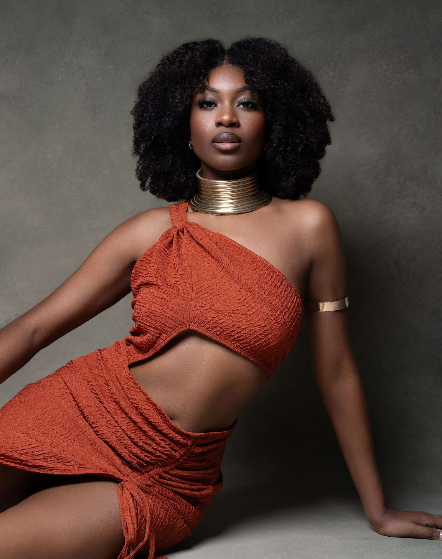 Woman in rust orange cut-out set with gold choker and cuff poses confidently against gray backdrop.