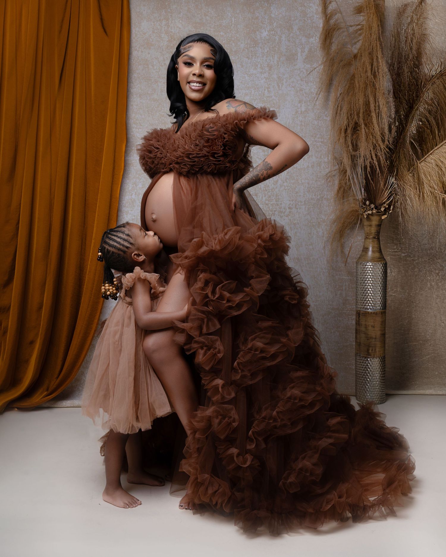 Pregnant woman in brown tulle maternity gown poses with young daughter in matching dress during studio photoshoot.