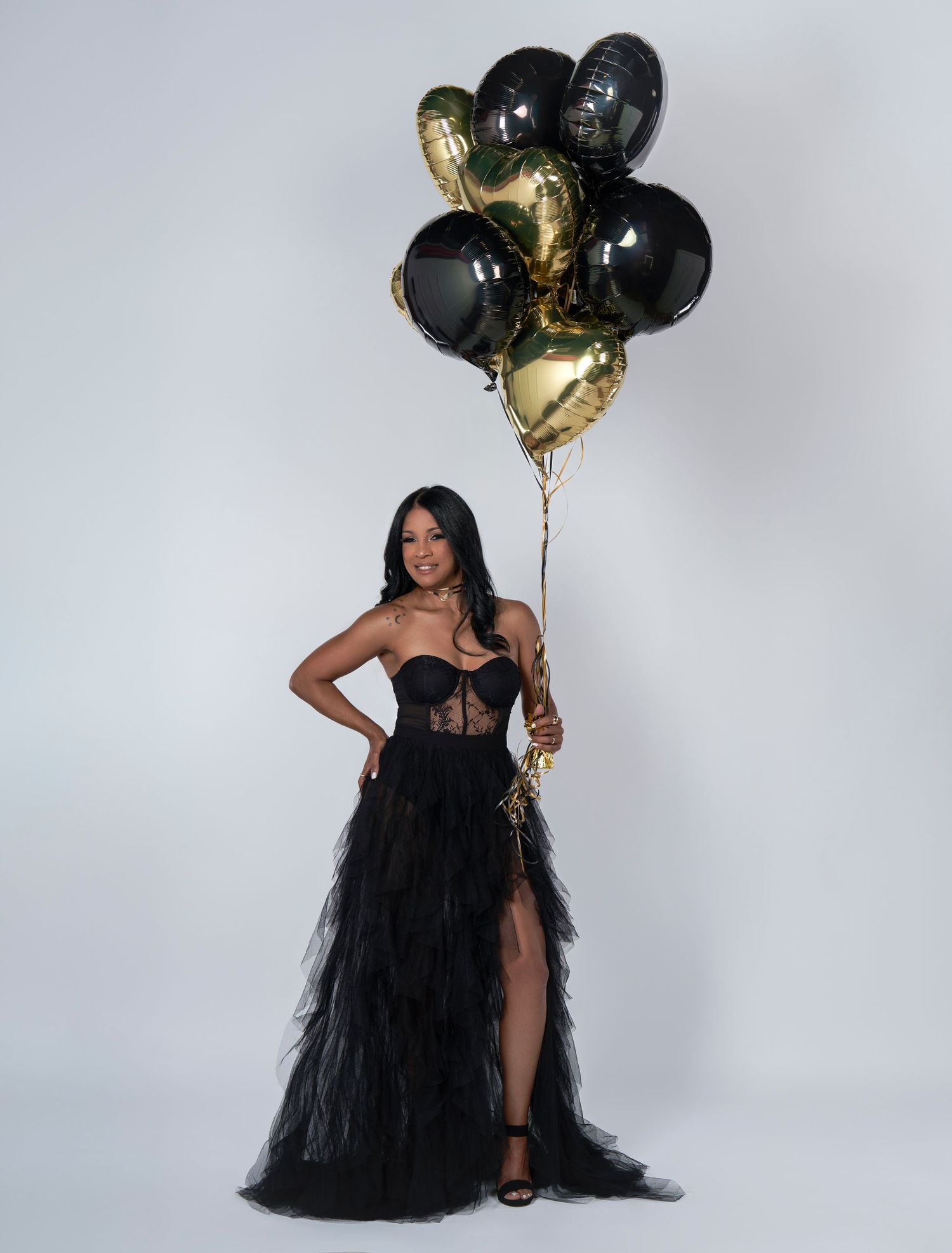 A person in an elegant black tulle gown with a corset bodice poses with black and gold balloons against a white backdrop.