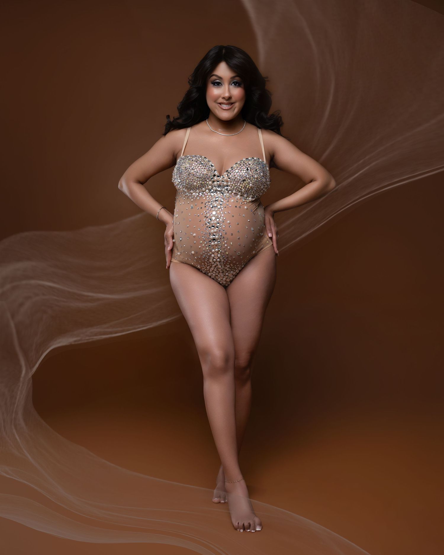 Pregnant woman in a bedazzled nude bodysuit poses confidently against a warm brown backdrop with flowing fabric.