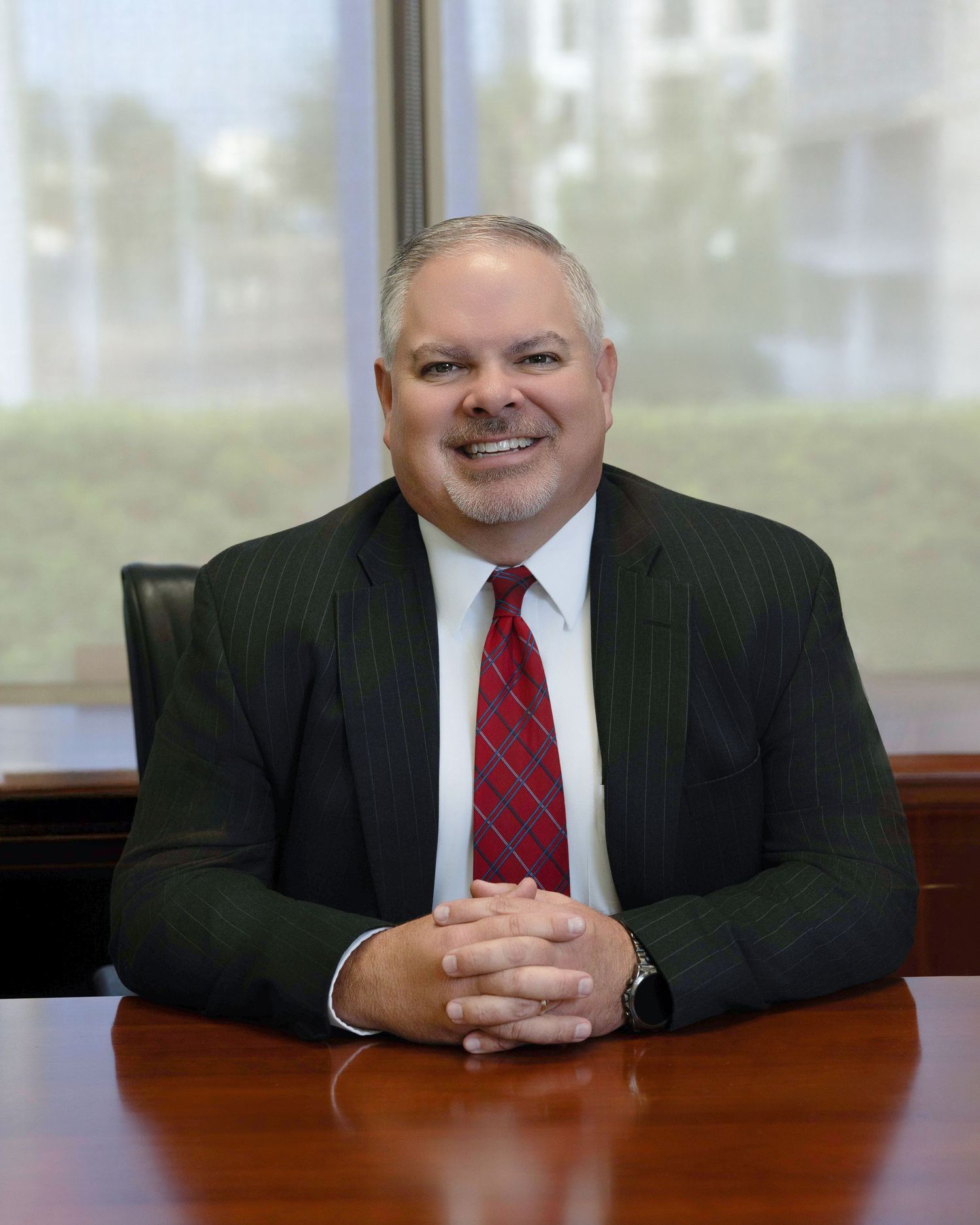Professional portrait of a person in a dark pinstripe suit and red tie sitting at a wooden desk in an office setting.