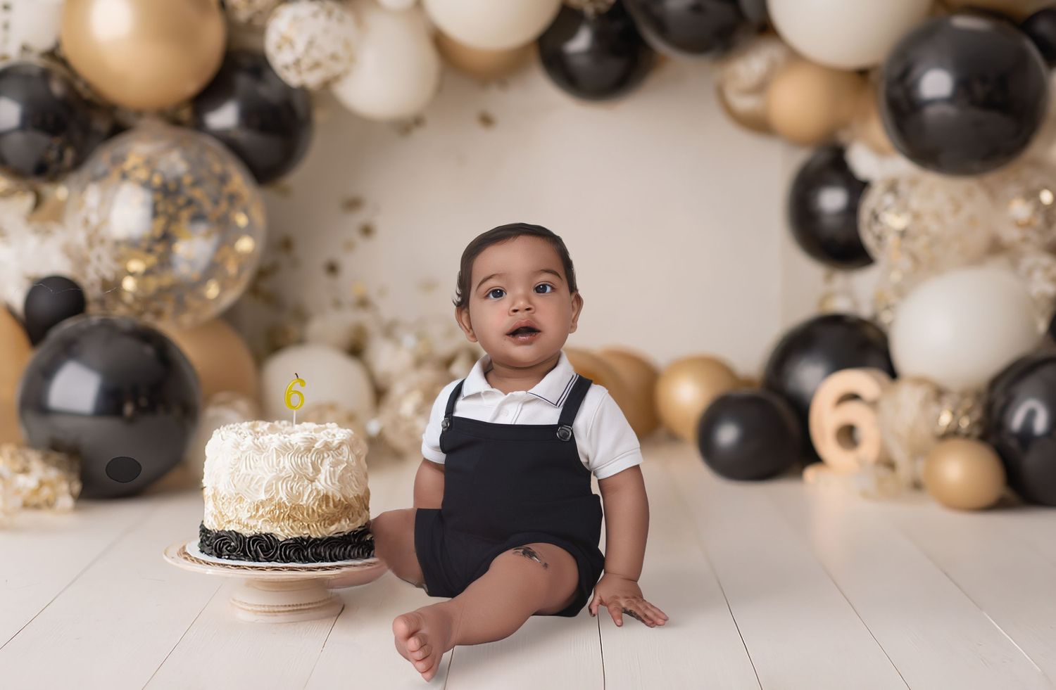 Baby boy in black romper sits before a smash cake, surrounded by black, gold, and white balloon arch.