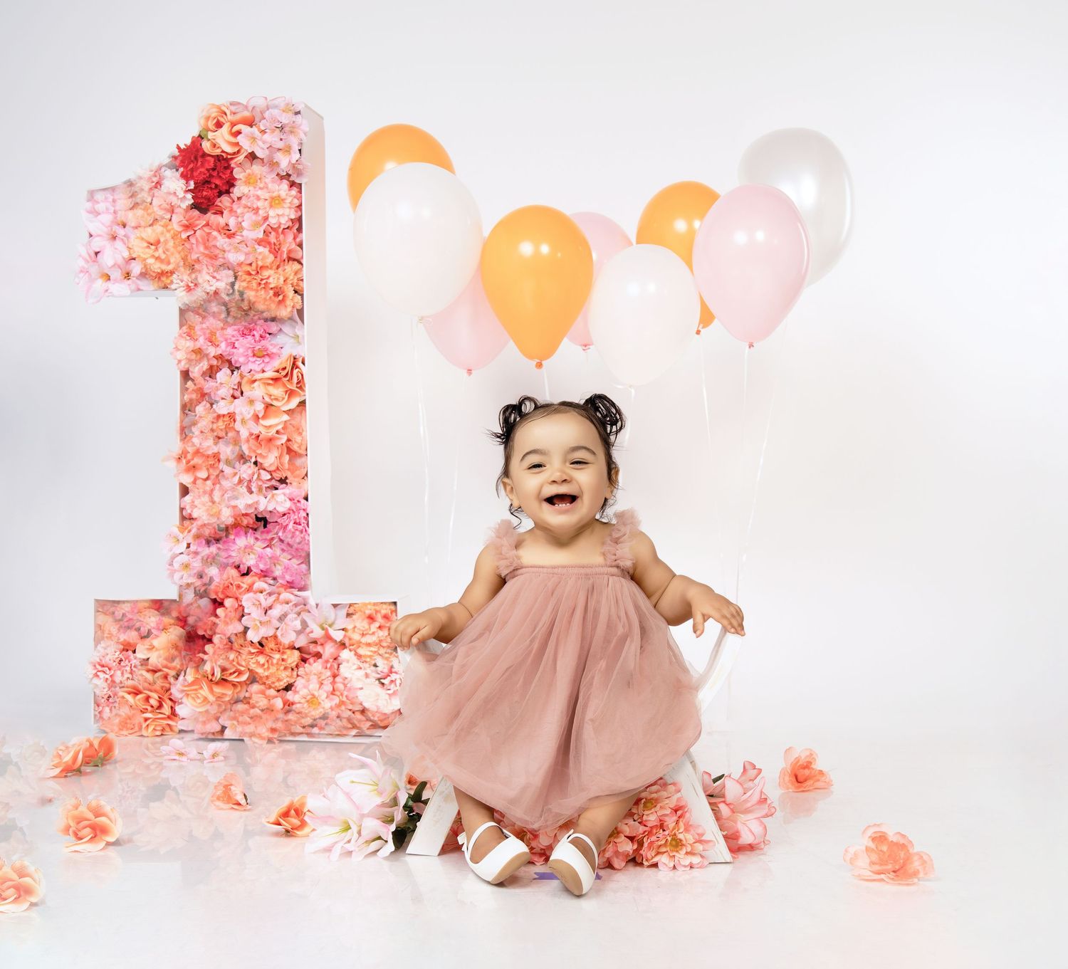 First birthday photo setup with pink floral wall, peach dress, and pastel balloons.
