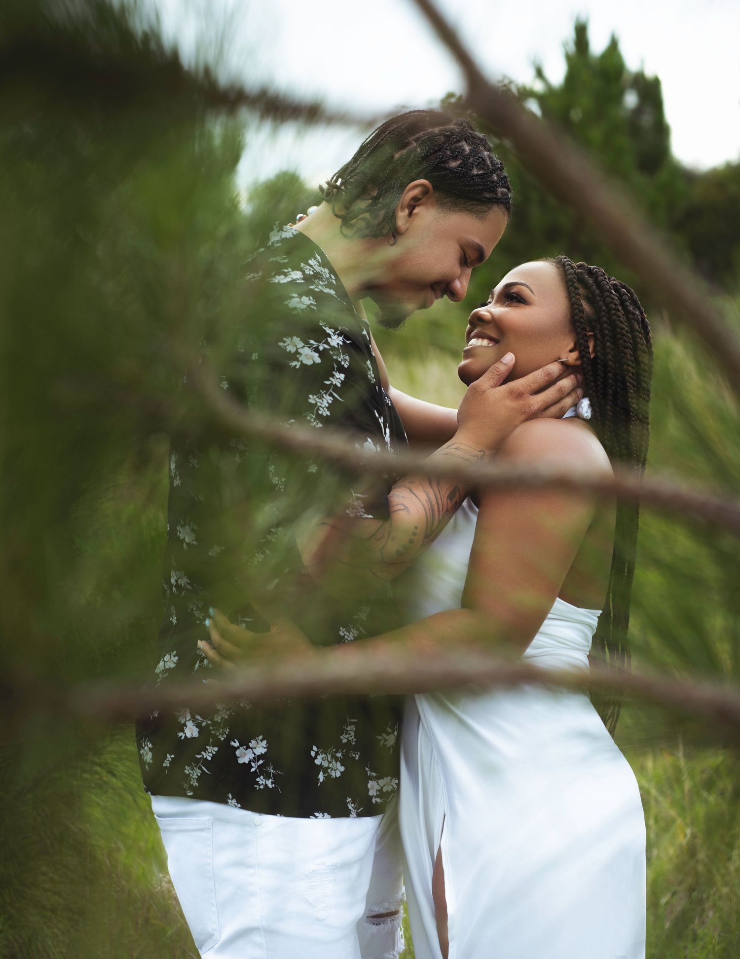 A romantic couple in white clothing share an intimate embrace outdoors among green foliage.