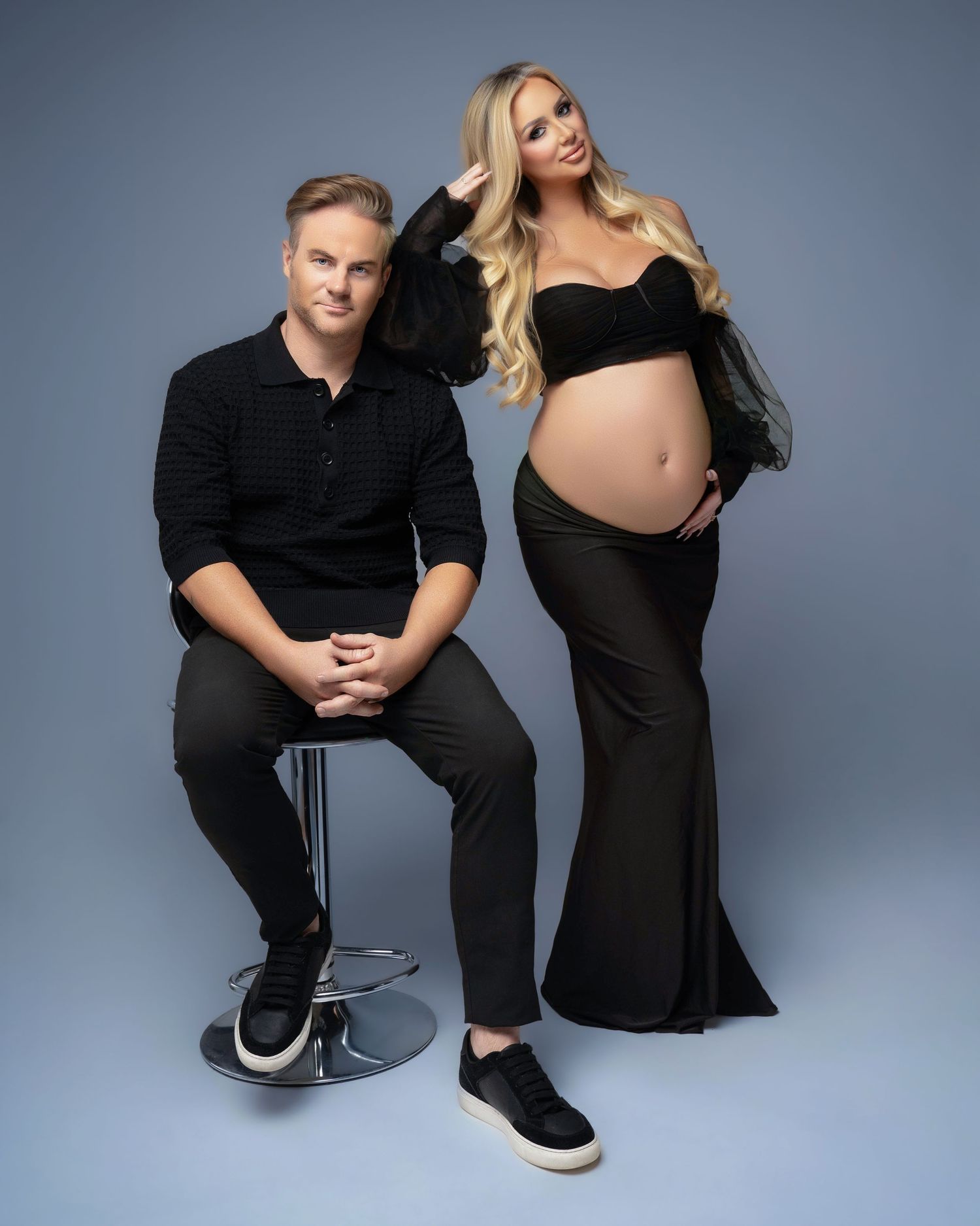Modern maternity studio portraits with couple in black outfits against blue backdrop.