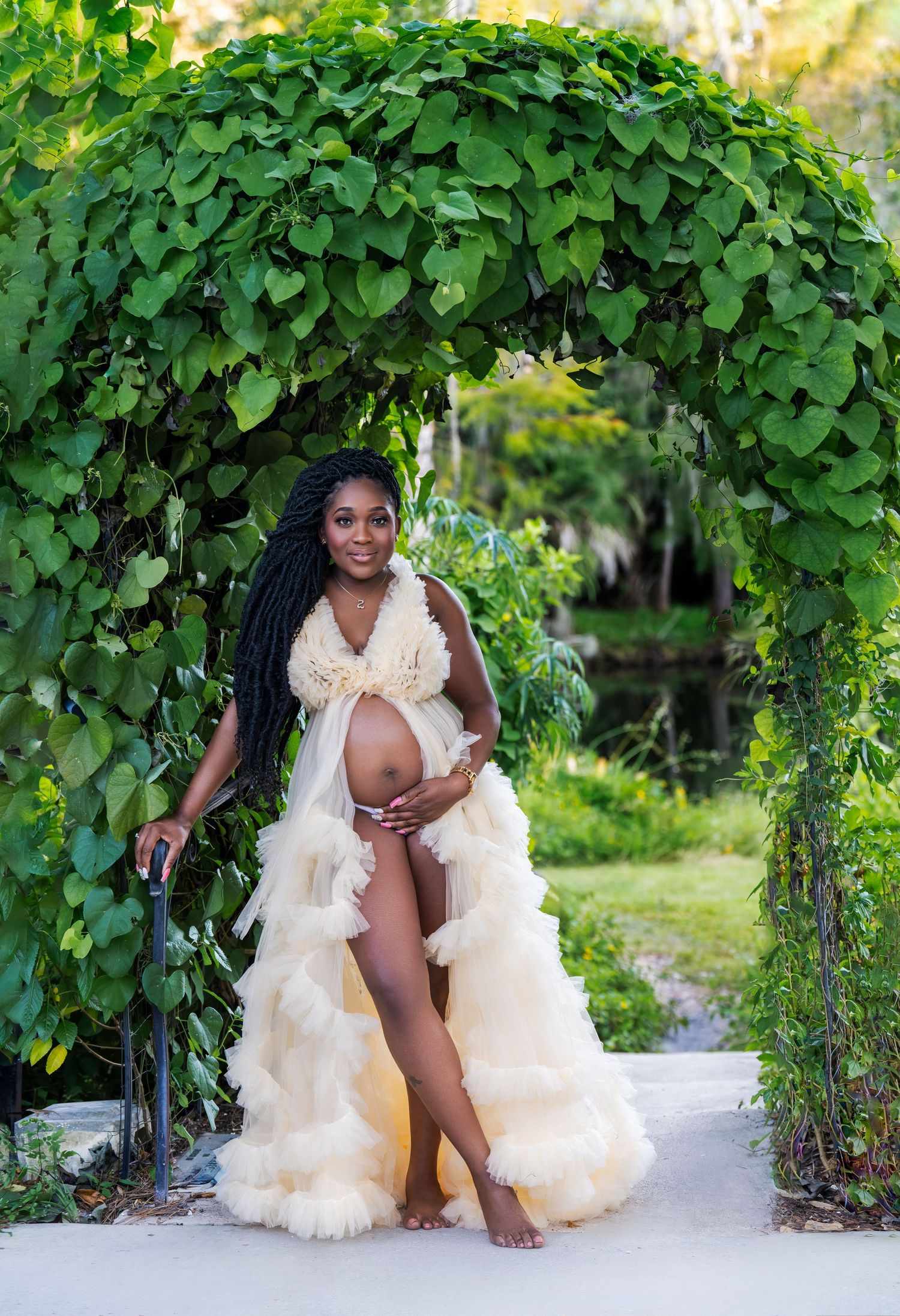 Outdoor maternity photography under green vine-covered archway with flowing white dress.
