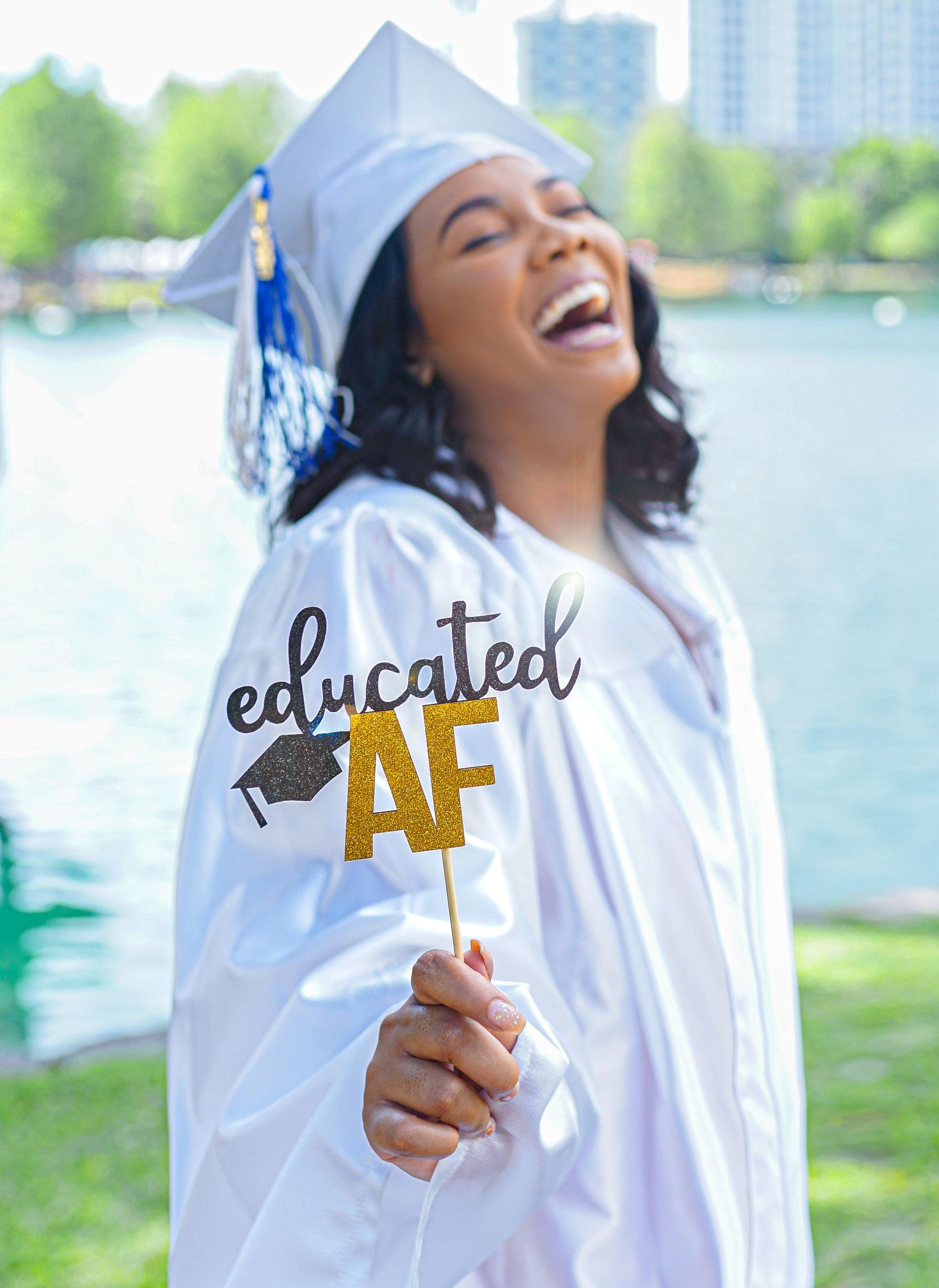 Graduate in white cap and gown holds up a celebratory sign reading 'educated AF' against a lakeside background.