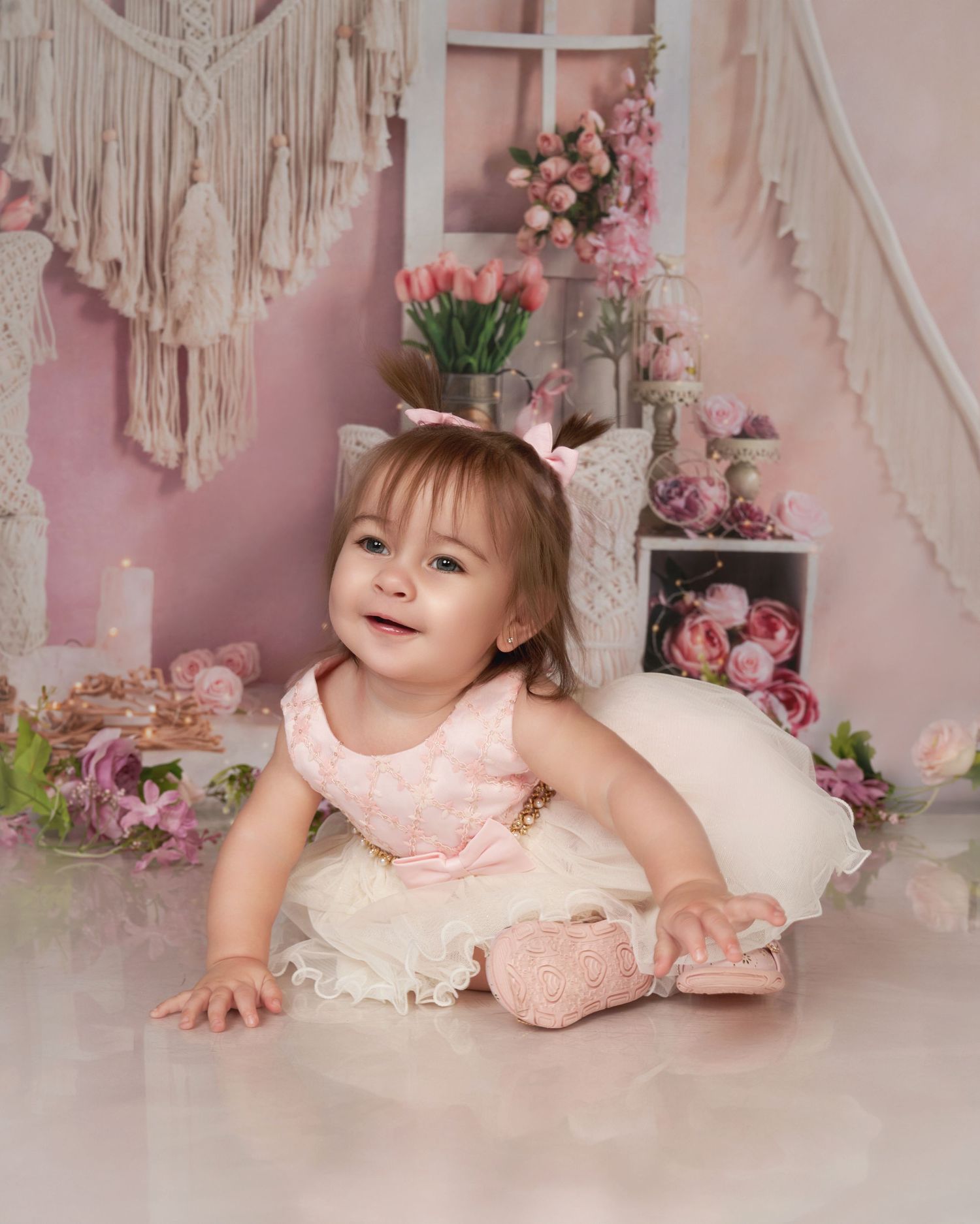 Baby in pink dress crawls on white floor with floral decorations and macrame wall hangings in pink nursery setting.